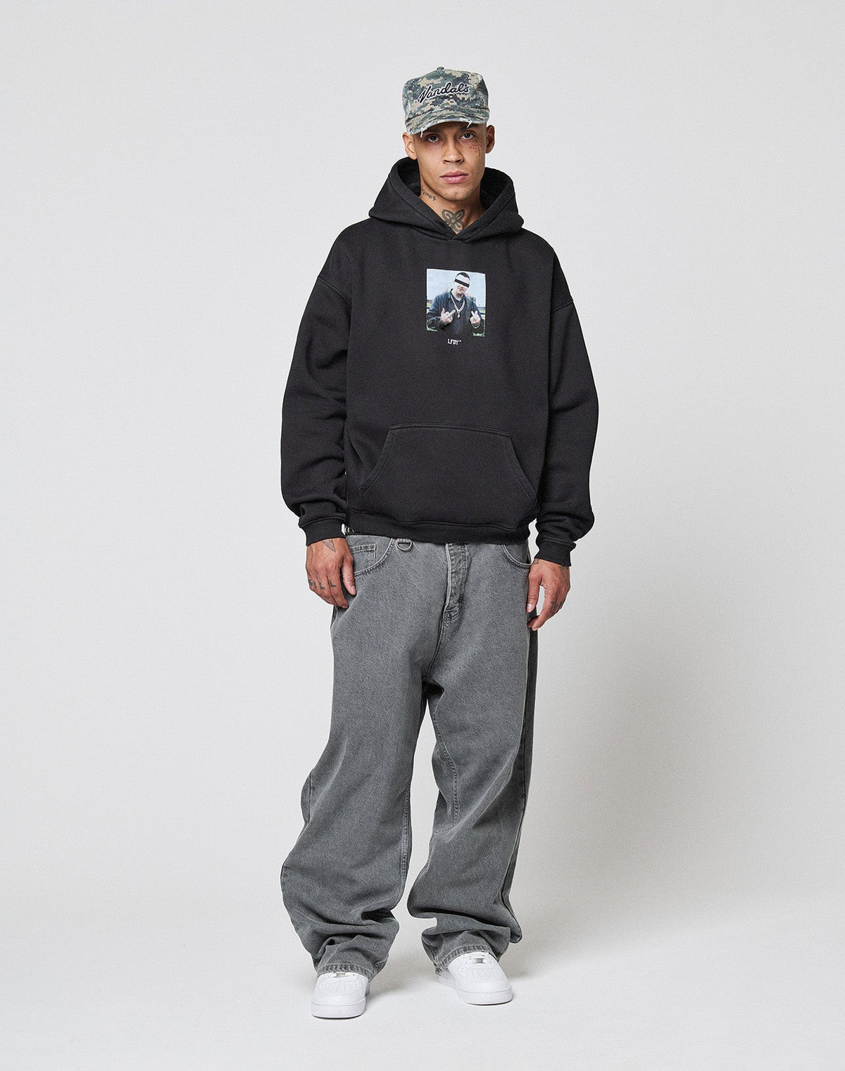 A person in a camo cap, LIVE FAST DIE YOUNG LFDY Gambling Hooded with front print, oversized baggy gray jeans, and white sneakers stands against a plain light background.