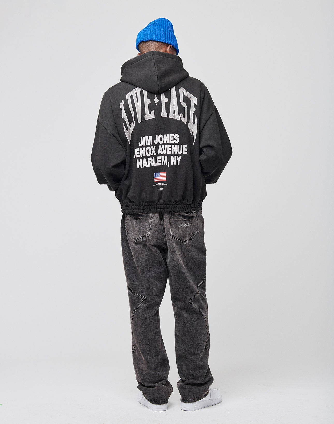 A person in an Oversize Fit black LIVE FAST DIE YOUNG Live Fast Lenox Hooded Zip hoodie with front and back prints, faded black jeans, white sneakers, and a blue beanie stands facing away against a plain light background.