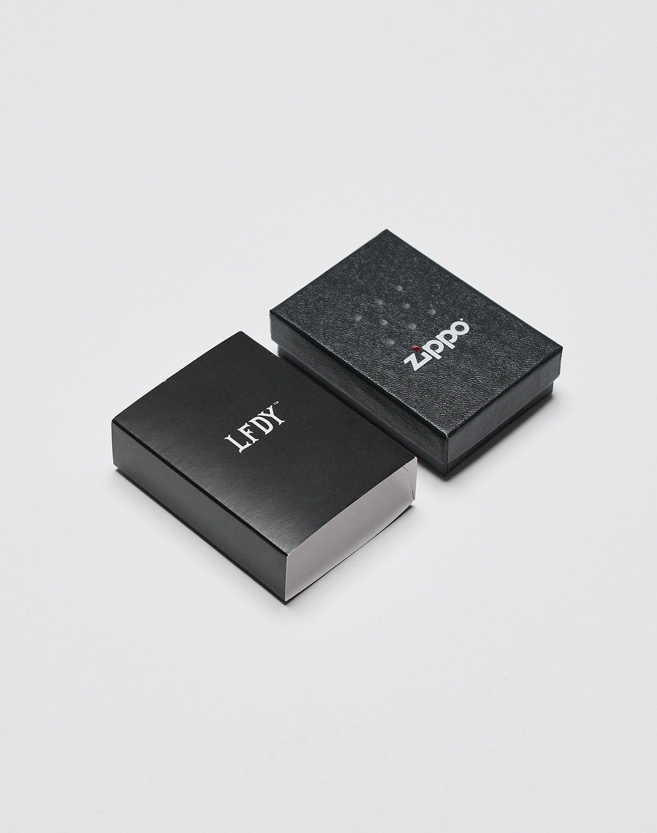 Two black rectangular boxes sit on a white surface; one closed with the Zippo logo, the other open showing LFDY x Zippo on its lid and a plain white interior, highlighting the LFDY x Zippo Limited Edition by LIVE FAST DIE YOUNG.