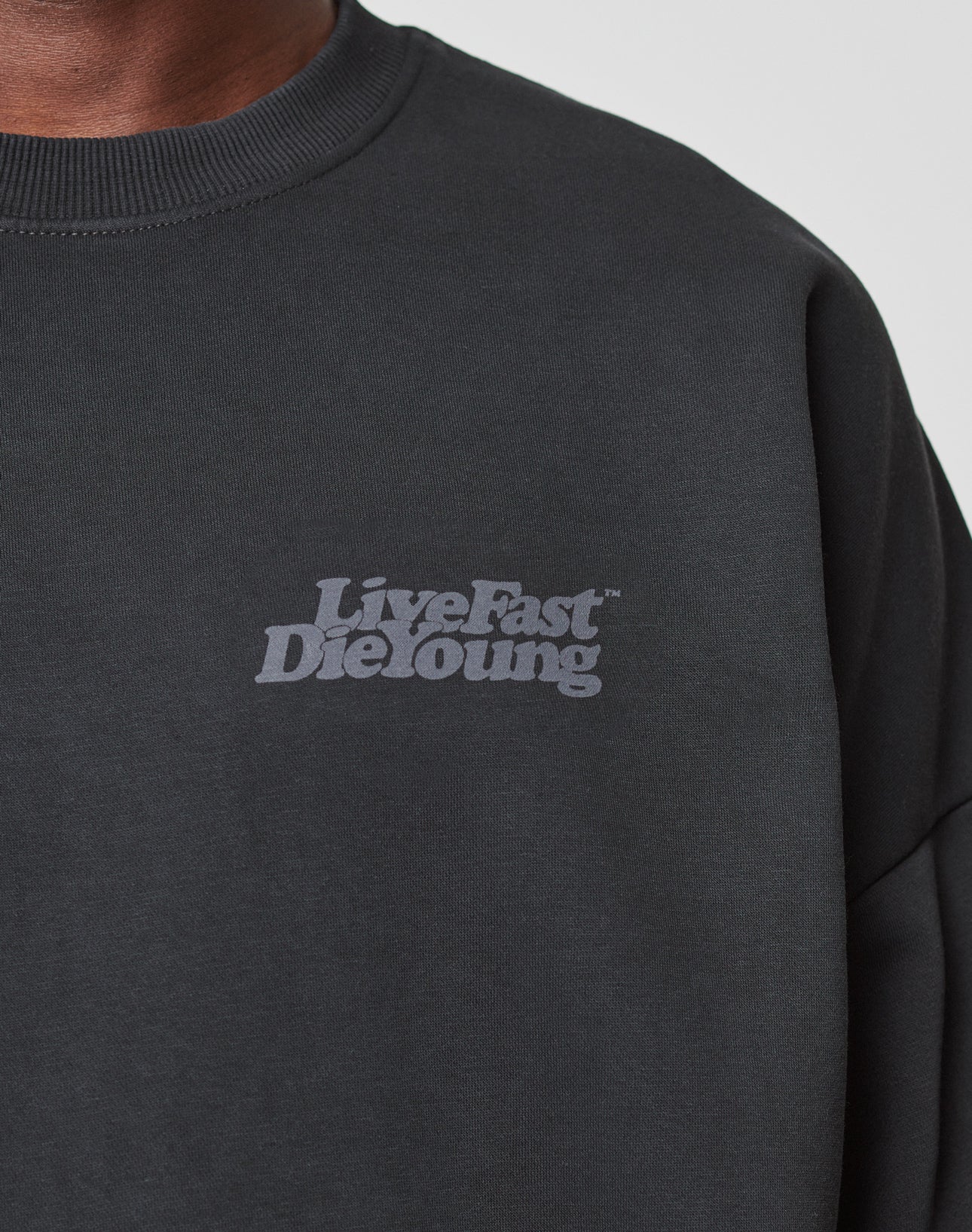 Close-up of a person in the Basic Acid Sweater by LIVE FAST DIE YOUNG—an oversize black sweatshirt featuring bold, gray “Live Fast Die Young” text on the left chest and made from a soft cotton-poly blend with vintage wash for retro style.