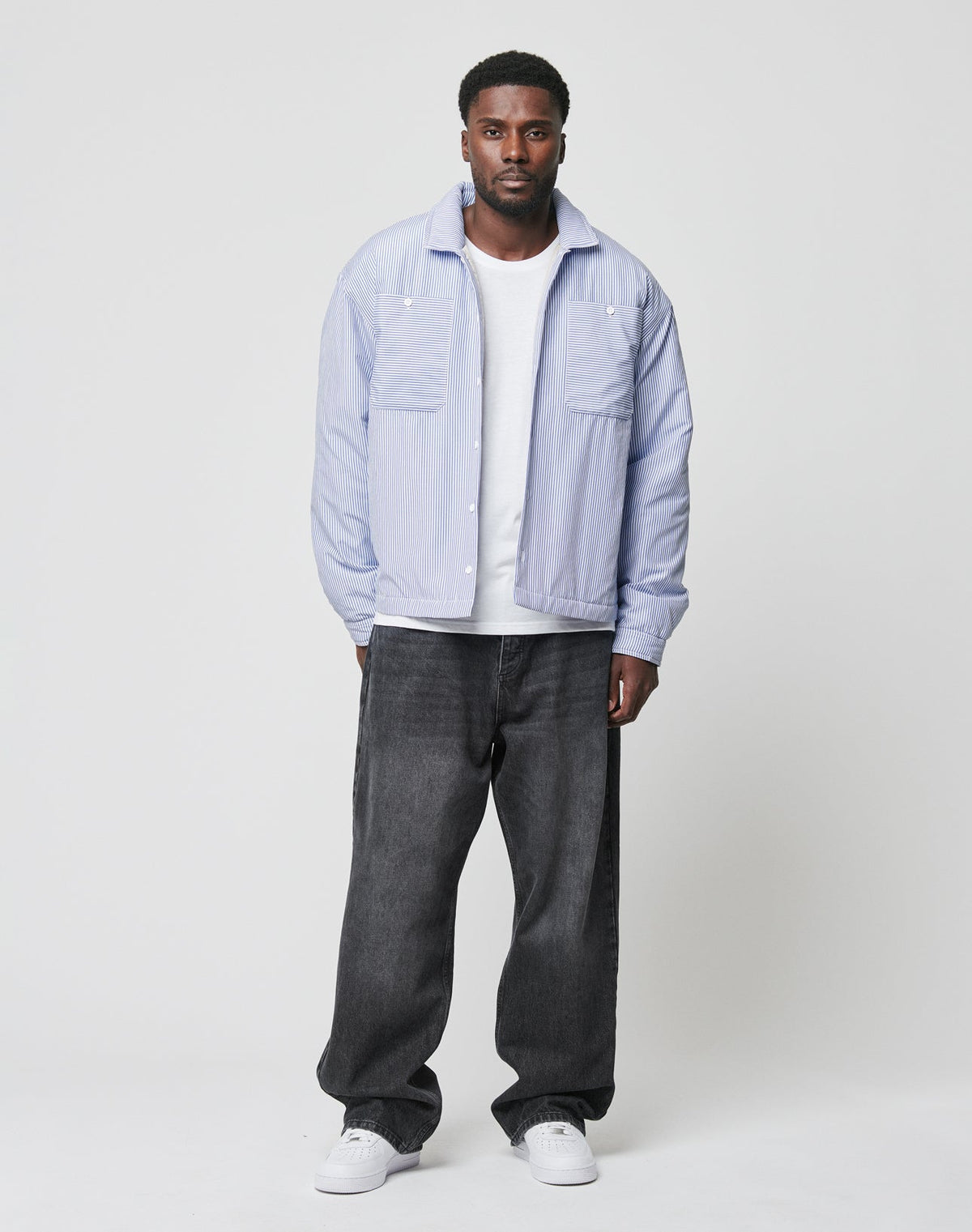 A man stands against a plain background in a light blue shirt over a white tee, wearing LIVE FAST DIE YOUNG’s LFDY Baggy Denim—black, 100% Baumwolle jeans with a wide straight leg—and white sneakers, looking directly at the camera with a neutral expression.