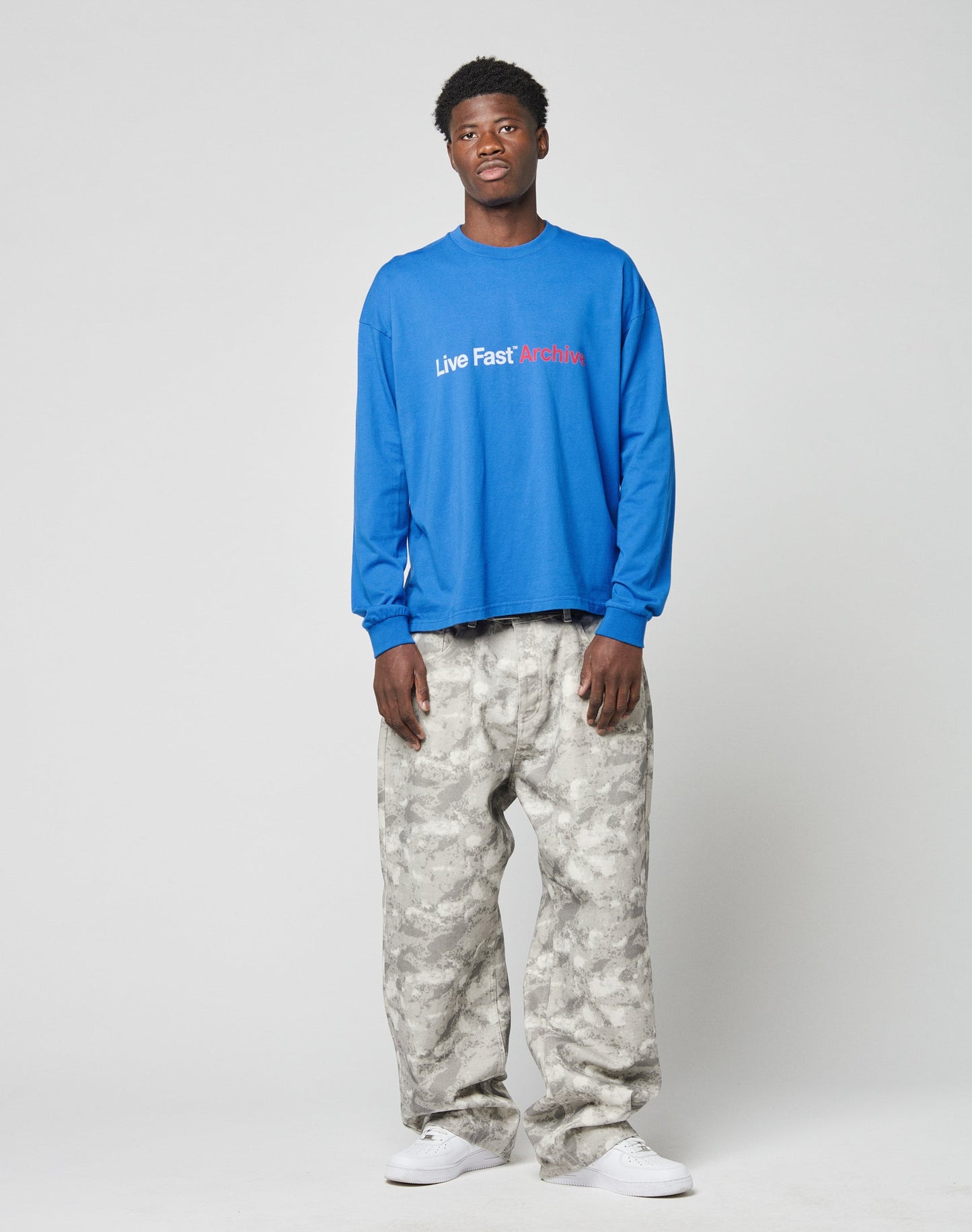 A man stands against a plain background wearing the LIVE FAST DIE YOUNG Live Fast Archive Longsleeve in blue with a front print, paired with loose light gray camo pants and white sneakers.