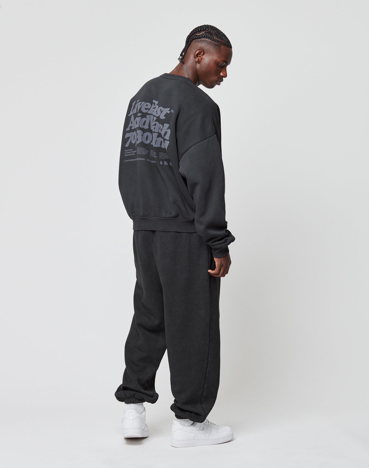 A person in an oversize fit LIVE FAST DIE YOUNG Basic Acid Sweater, featuring large gray text on the back, with matching black sweatpants and white sneakers, stands against a plain white background facing slightly away from the camera.