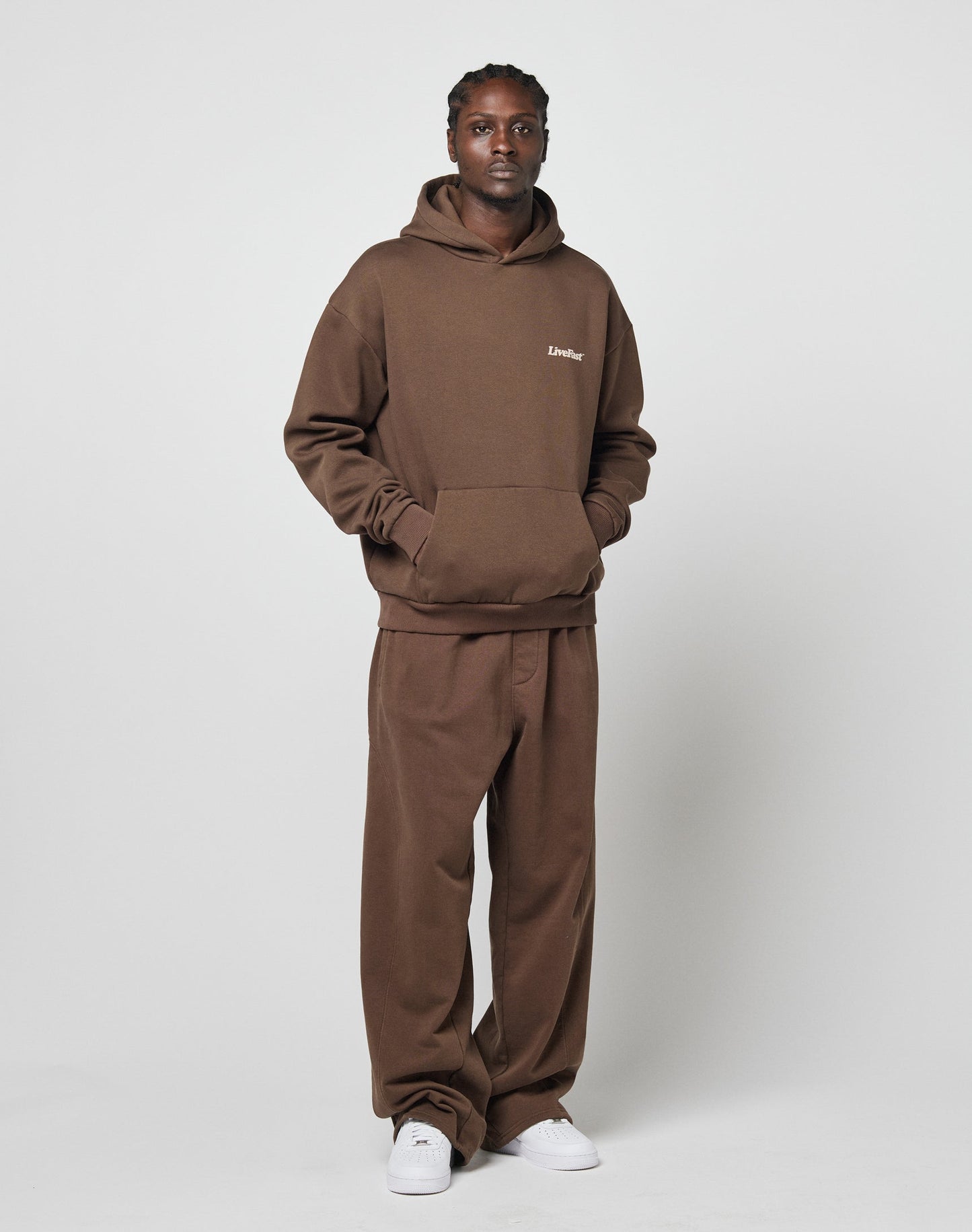 A man wearing the LIVE FAST DIE YOUNG Basic Wide Jogger in brown, featuring a wide fit with breiter Elastikbund and seitliche Eingrifftaschen, stands against a white background with his hands in his hoodie pocket, finished with white sneakers.