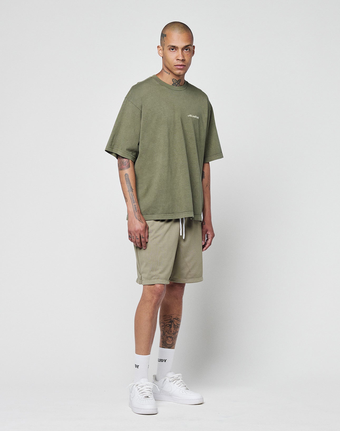 A person with tattoos on their arms, neck, and legs stands against a plain white background wearing the LIVE FAST DIE YOUNG Basic +49 Chest Tee in olive green, matching shorts, white crew socks, and sneakers.