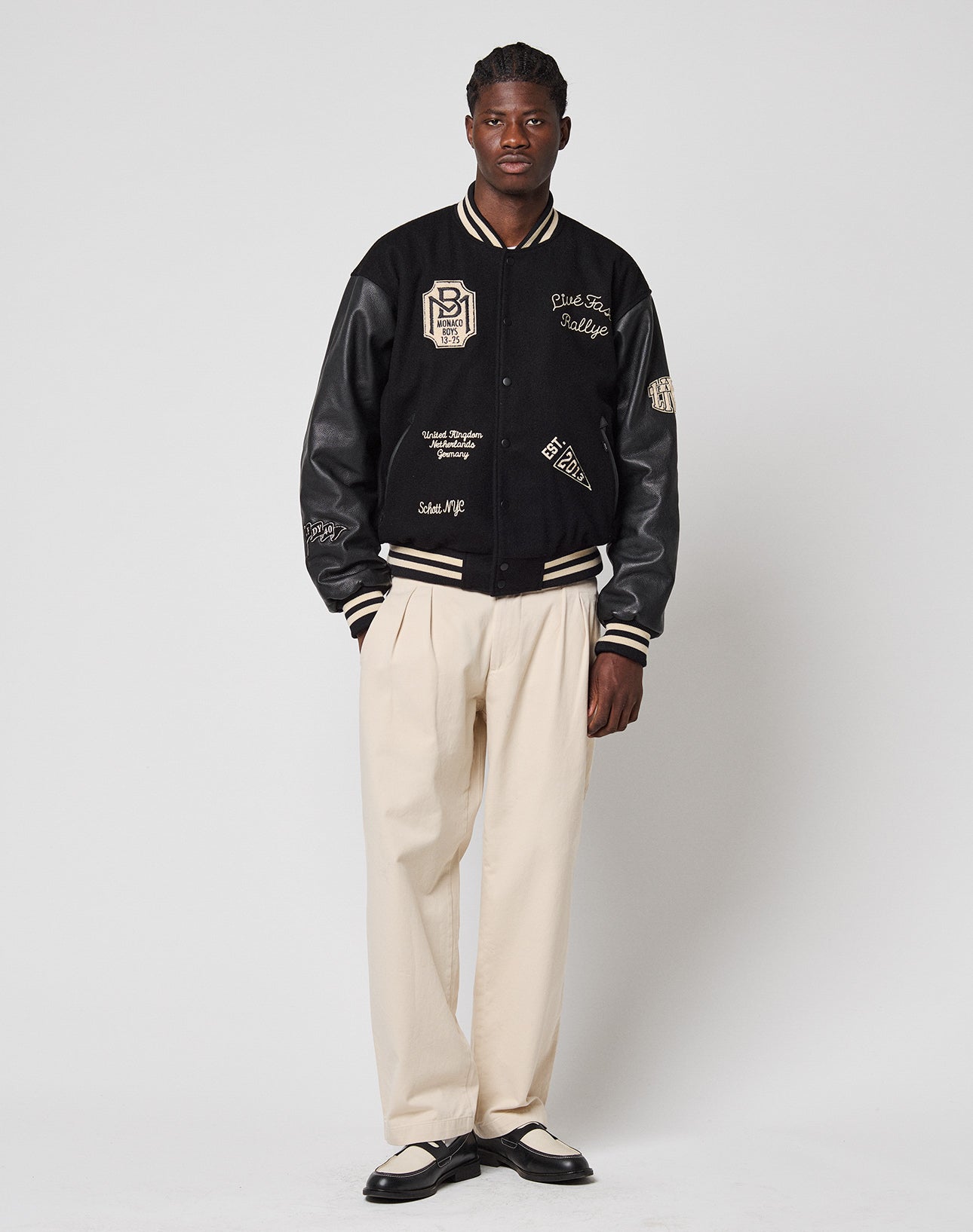 A man stands against a white background wearing a black varsity jacket with cream details, LIVE FAST DIE YOUNG Wide Chino trousers in 100% cotton, and black-and-white dress shoes. He faces forward neutrally, hands at his sides.