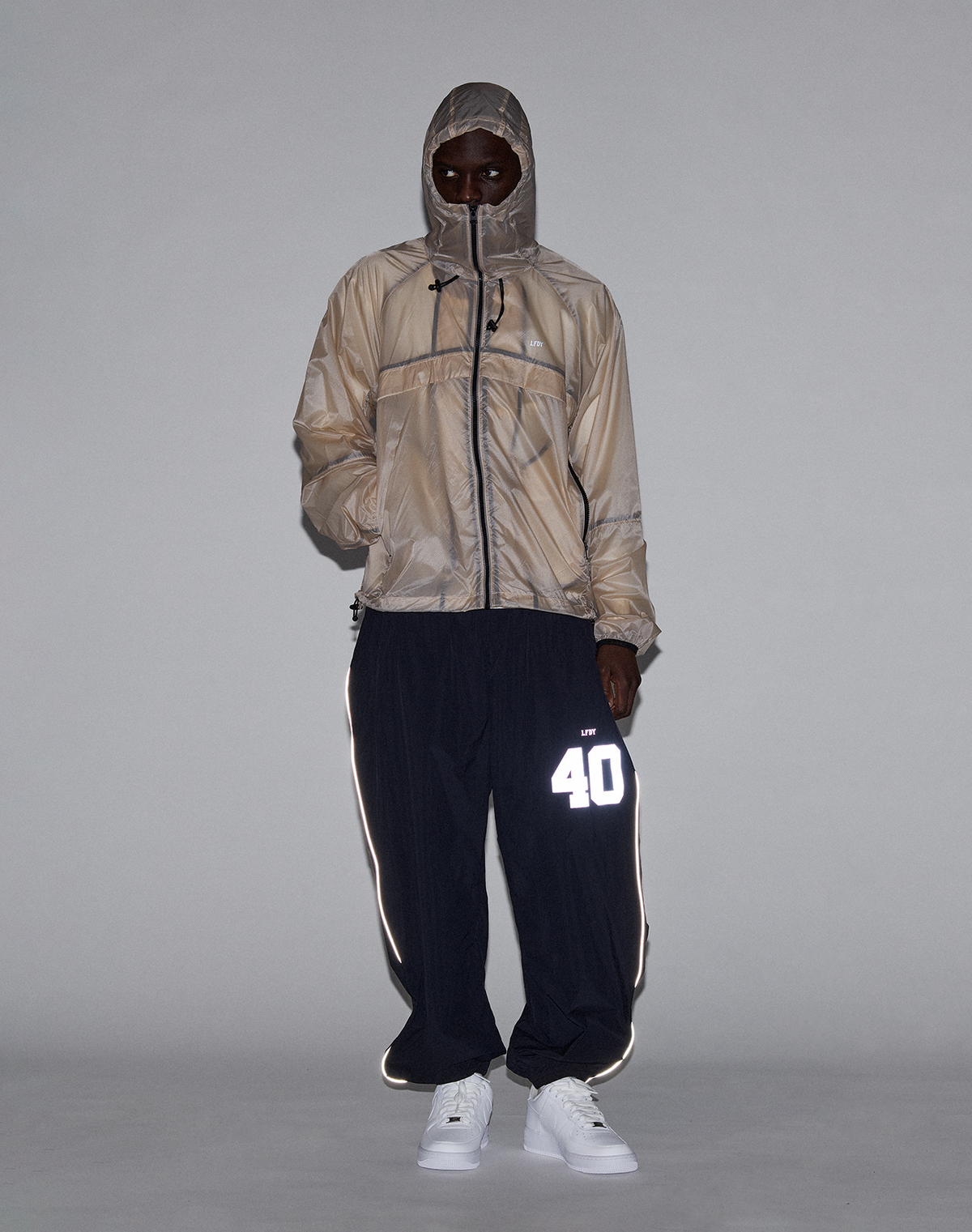 A person in a beige hooded jacket with zippered face cover, white sneakers, and black LIVE FAST DIE YOUNG LFDY Performance Trackpants featuring a reflective “40” print on the left leg stands against a plain gray background.