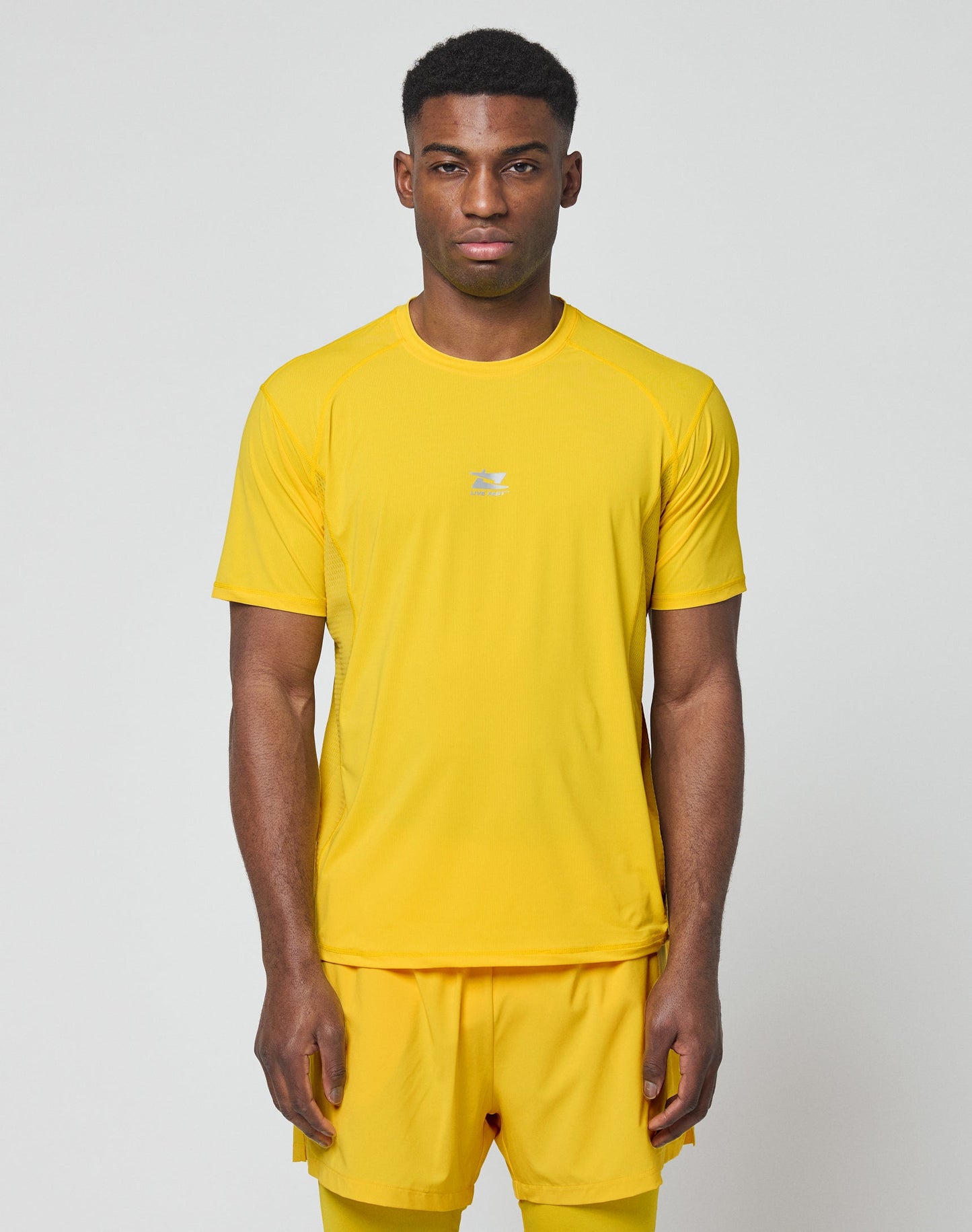 A man faces forward in the LIVE FAST DIE YOUNG Live Fast Performance Tee and matching shorts, slim fit, bright yellow with reflective print, set against a plain light gray background.