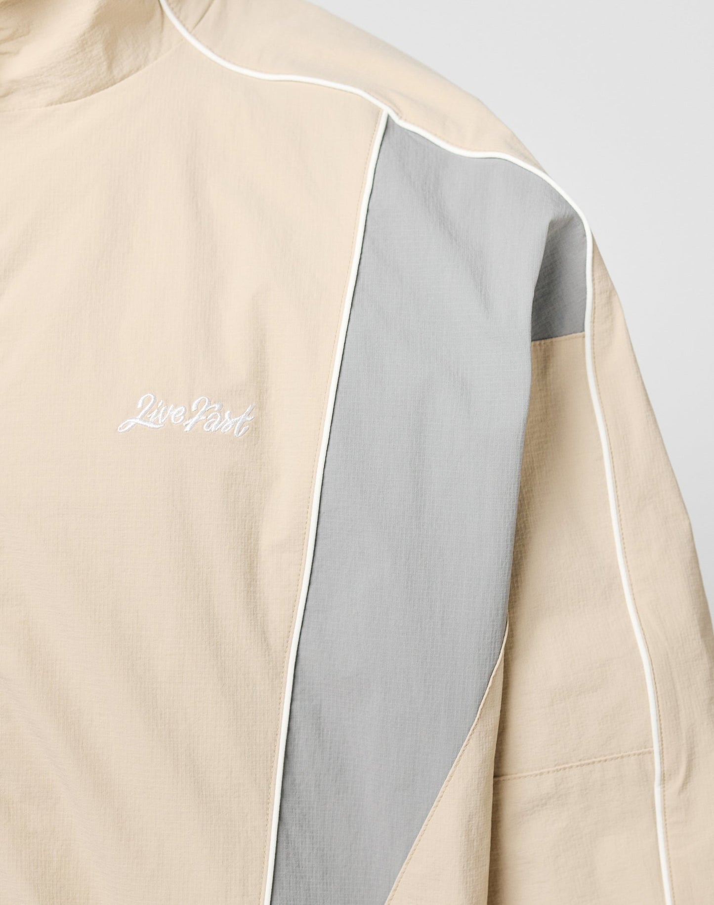 The Basic Nylon Trackjacket by LIVE FAST DIE YOUNG features a beige and light gray color block design, white piping, Live Fast embroidery on the left chest, regular fit, high collar, and is crafted from nylon and polyester.