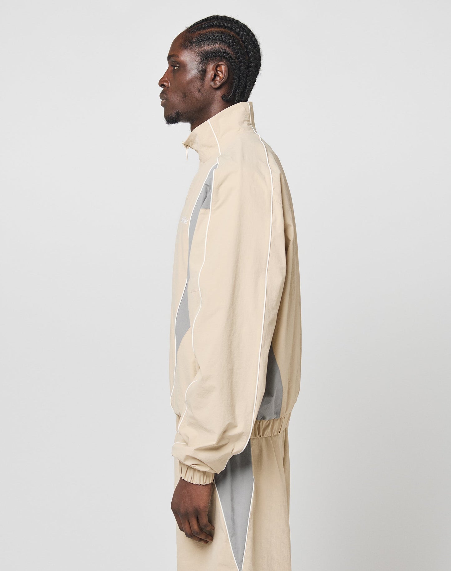 A man with braided hair stands in profile, wearing the LIVE FAST DIE YOUNG Basic Nylon Trackjacket and matching pants in regular fit, set against a plain light gray background.