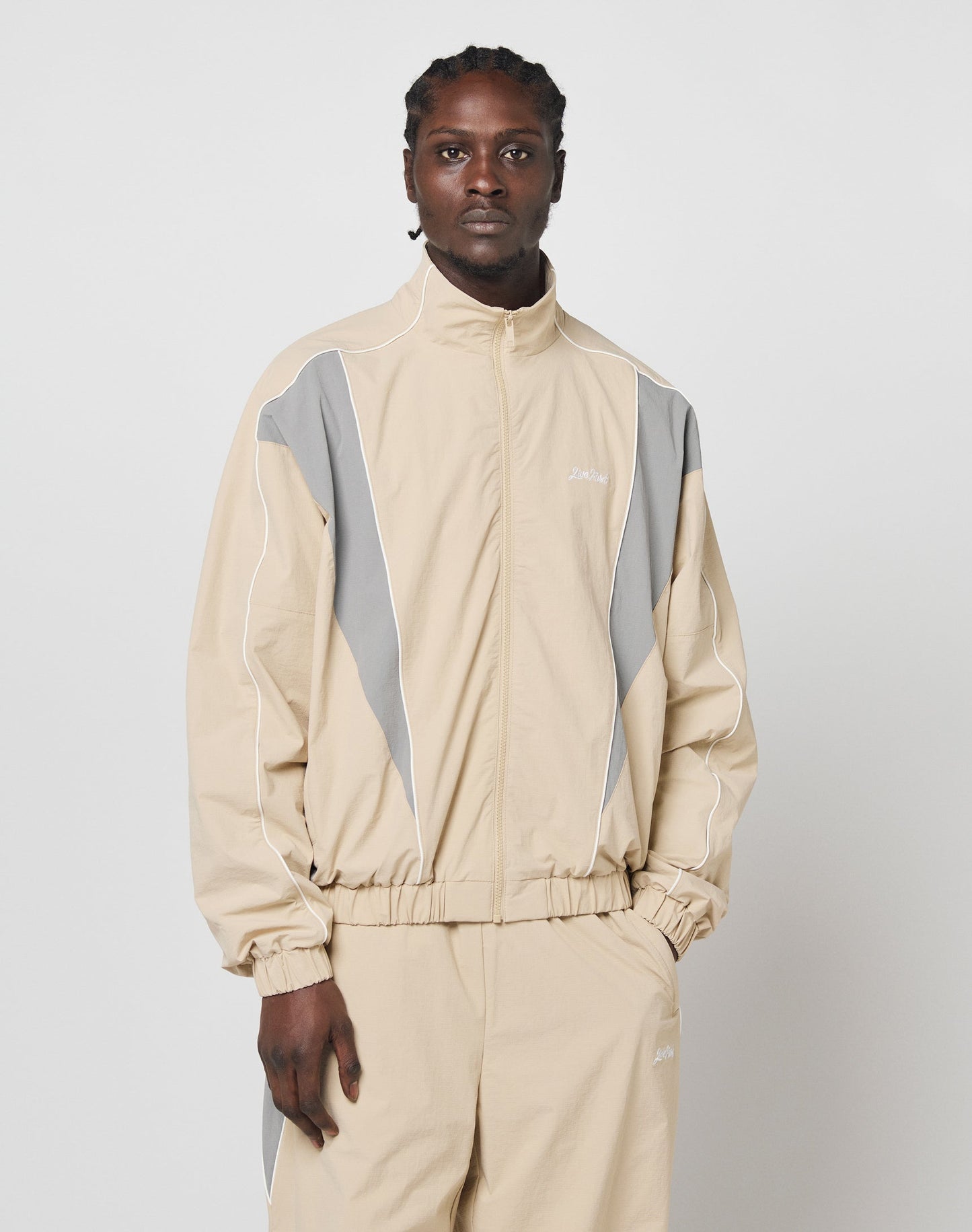 A person models the LIVE FAST DIE YOUNG Basic Nylon Trackjacket in beige with gray and white accents, standing against a plain white background and looking at the camera with a neutral expression.
