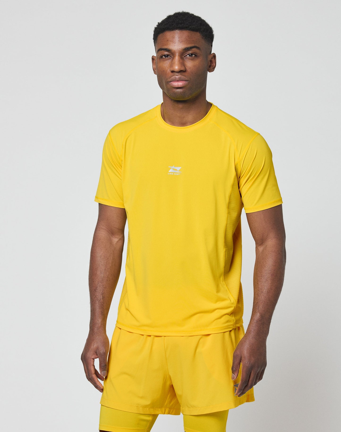 A man stands against a plain background wearing the LIVE FAST DIE YOUNG Live Fast Performance Tee and matching shorts in bright yellow nylon spandex with a reflective print. He looks forward neutrally, arms relaxed at his sides.