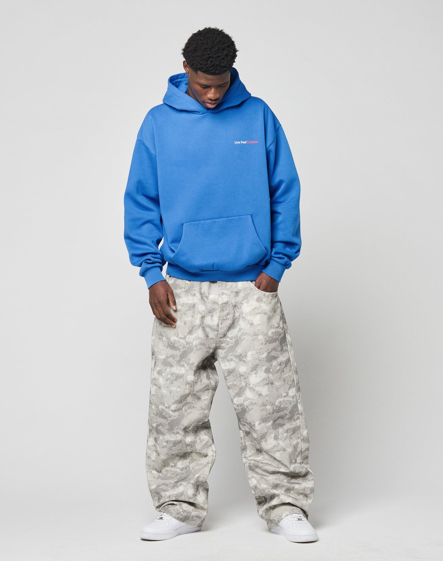 A man stands against a plain background wearing the LIVE FAST DIE YOUNG LFDY F\*ck Phones Hooded in bright blue, paired with light gray camo baggy pants and white sneakers, looking down with his hands in his pockets.