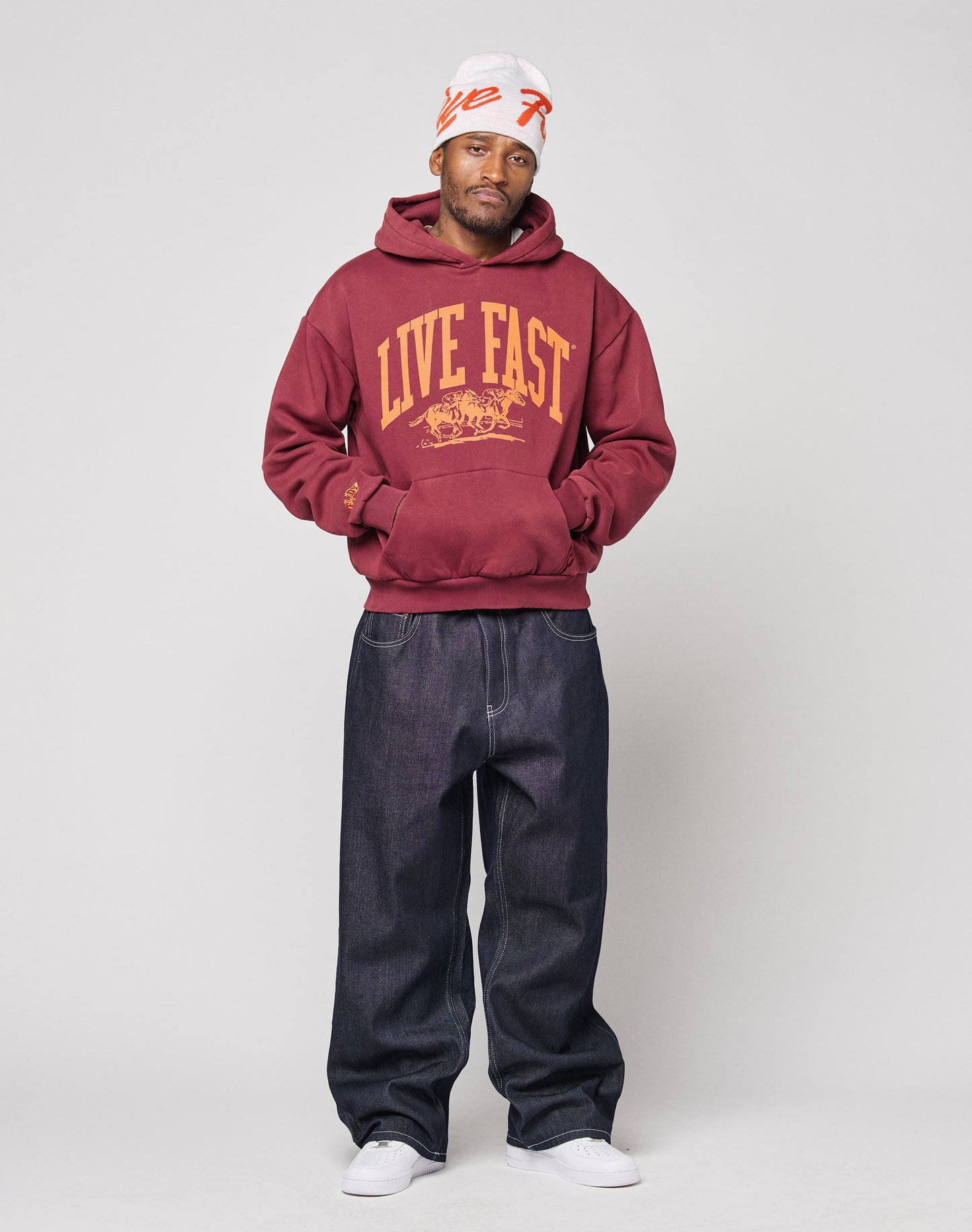 A man wears a white beanie, loose dark jeans, white sneakers, and the LIVE FAST DIE YOUNG “Live Fast Stallion Hooded” oversize maroon hoodie with “LIVE FAST” text, standing against a plain light background.