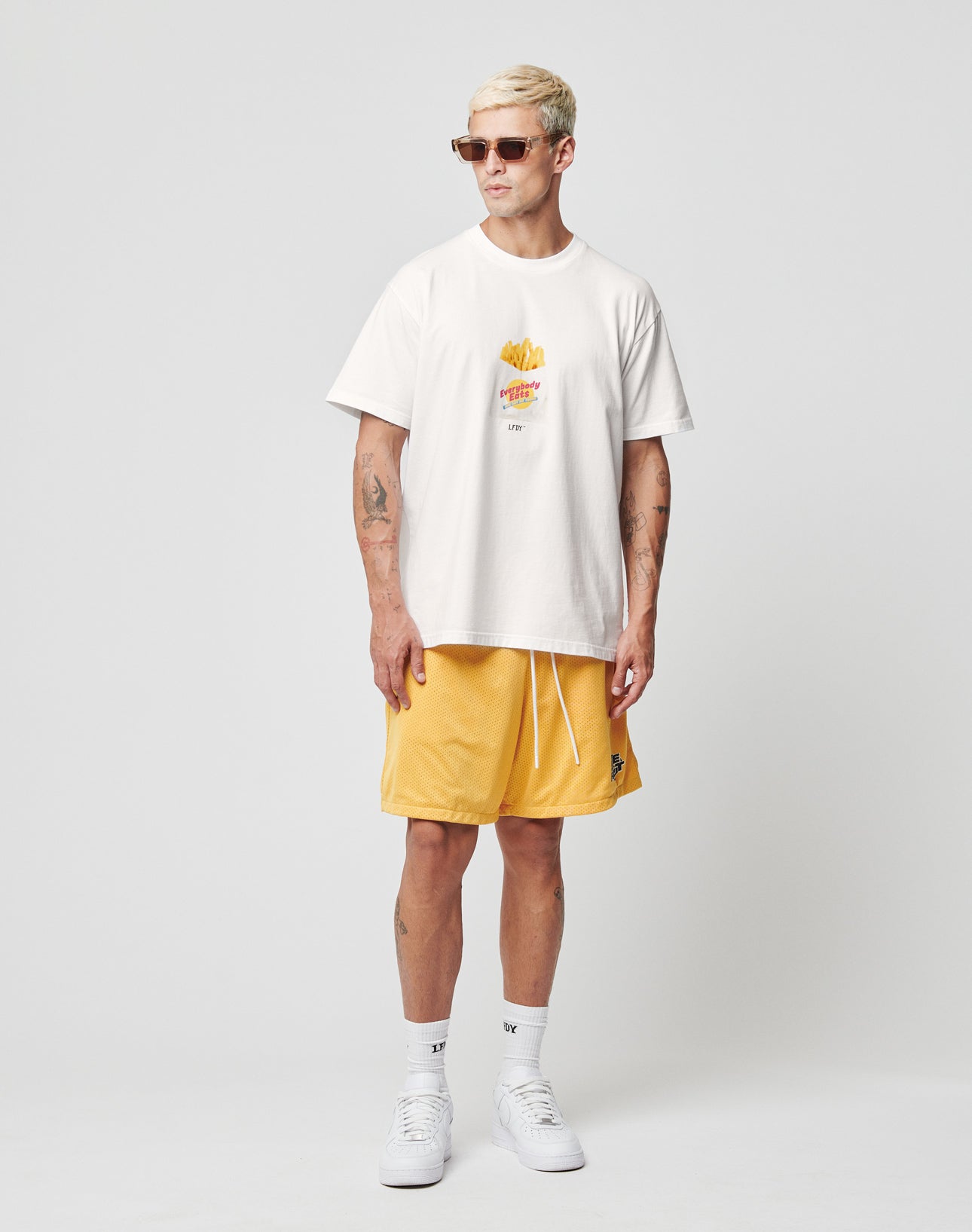 A person with short blond hair wears sunglasses, yellow shorts, white socks, white sneakers, and a LIVE FAST DIE YOUNG LFDY Fries Tee featuring a small fries graphic, standing against a plain light gray background.