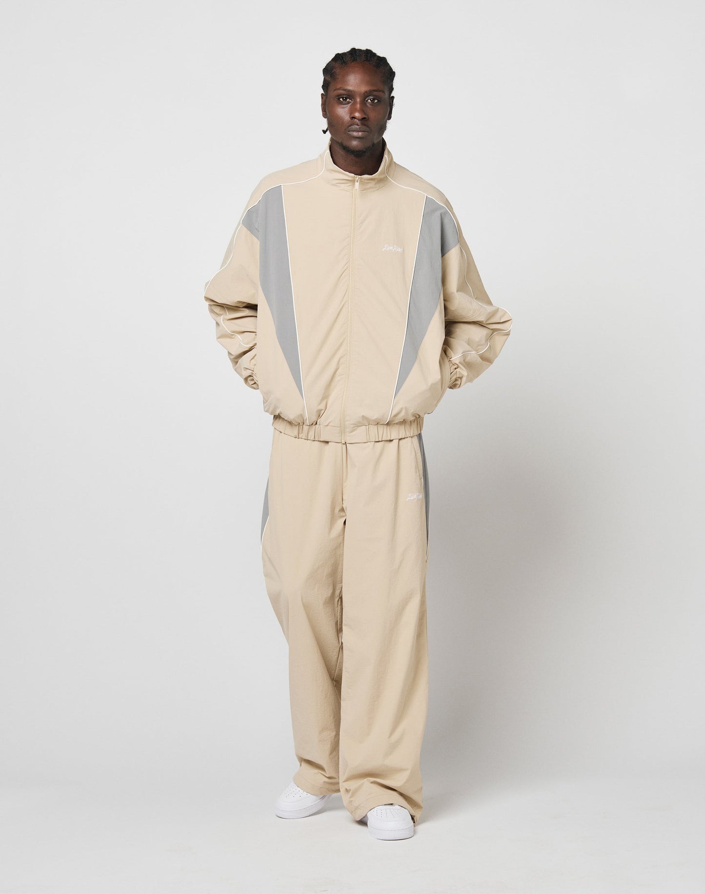 A man with braided hair stands against a white background wearing LIVE FAST DIE YOUNG Basic Nylon Trackpants in beige and gray wide fit, featuring Reißverschluss Seitentaschen and an Elastik-Kordelzug, paired with white sneakers. Hands in jacket pockets.