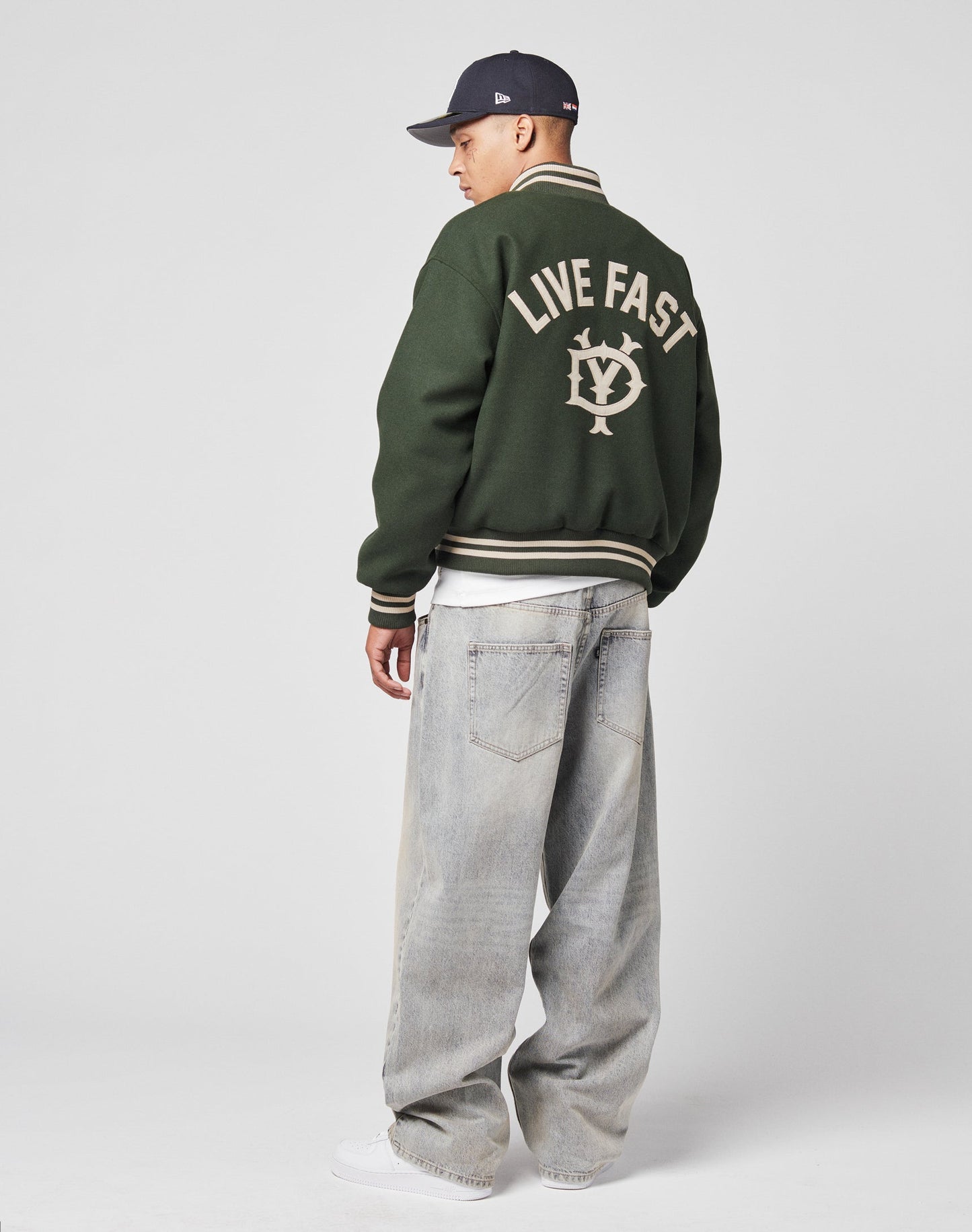 A person wears the LIVE FAST DIE YOUNG Live Fast College Jacket with Rückenstick and a circular back logo, loose light-wash jeans, white sneakers, and a dark baseball cap against a plain light background.