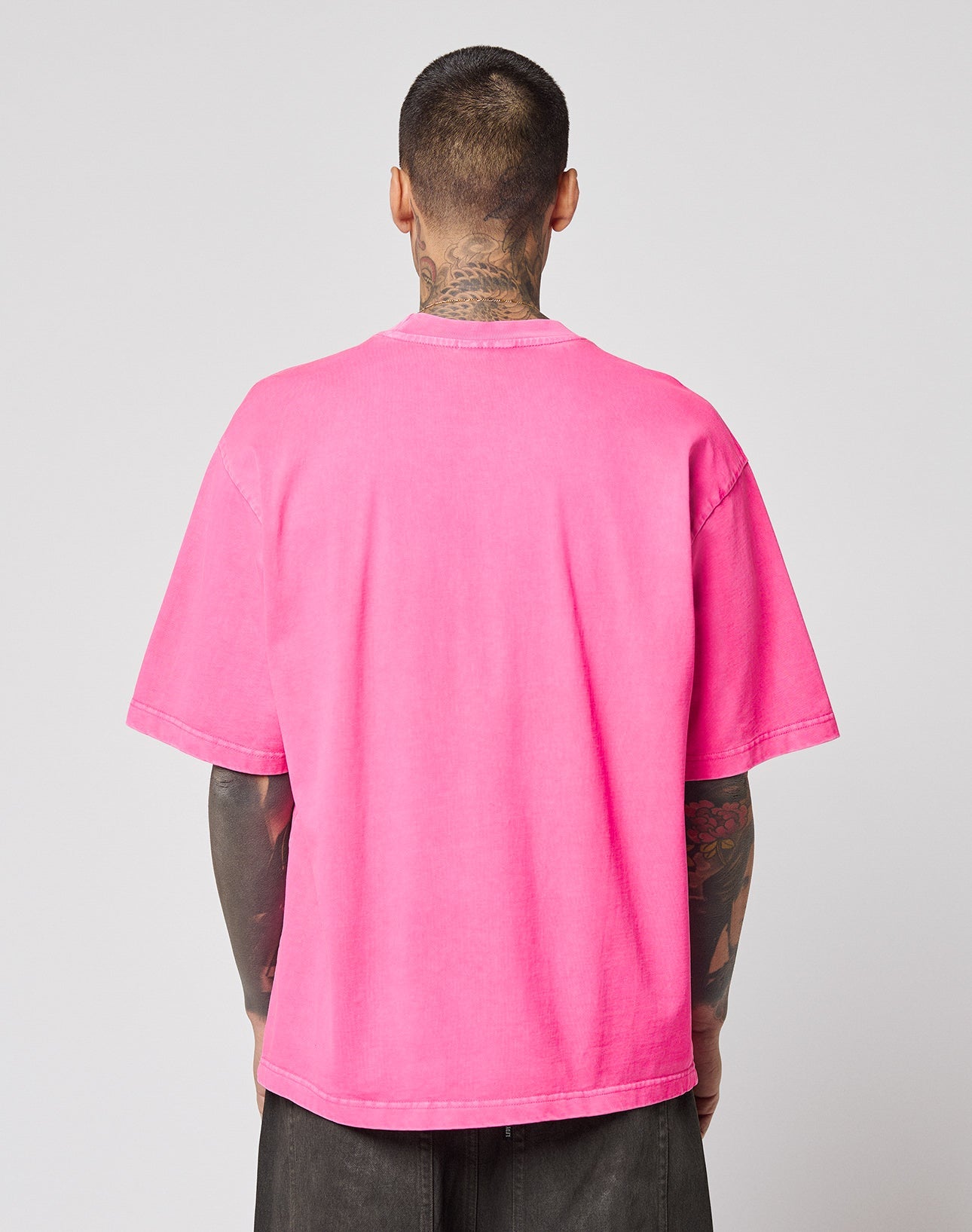 A person with short hair and visible tattoos on their neck and arms is shown from the back, wearing the bright pink LFDY Triple Racing Tee by LIVE FAST DIE YOUNG, styled with dark pants against a plain light background.