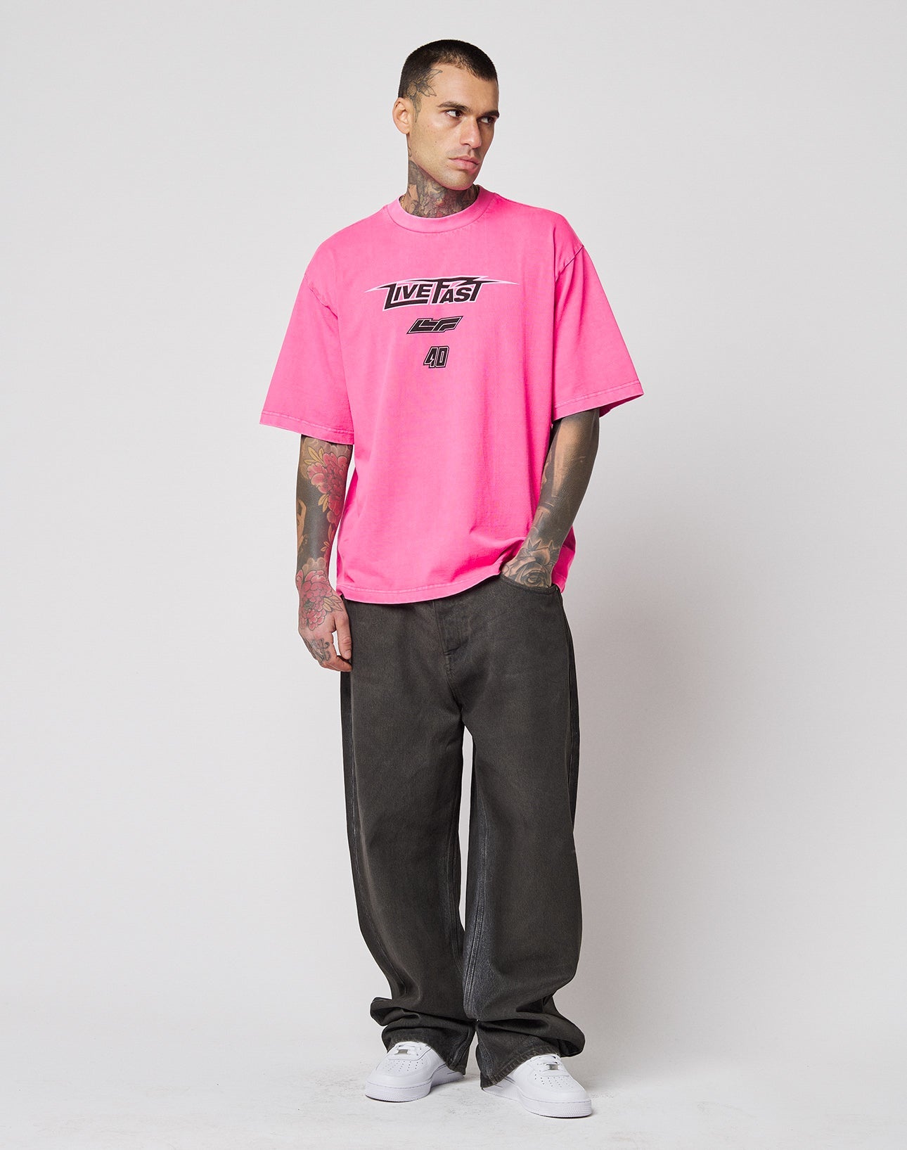 A tattooed man stands against a plain background in the LIVE FAST DIE YOUNG LFDY Triple Racing Tee, an oversize fit with multilogo frontprint, paired with loose dark pants and white sneakers, looking slightly to the side with hands in his pockets.