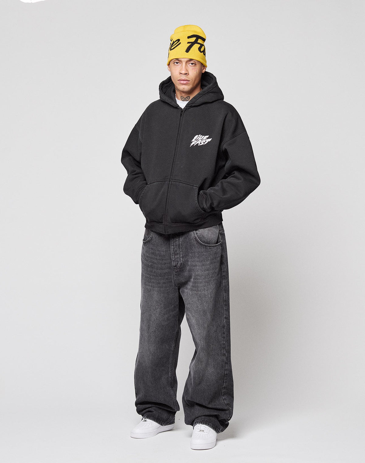 A person stands against a plain background wearing a LIVE FAST DIE YOUNG LFDY Logo Hooded Zip in black, loose dark jeans, white sneakers, and a yellow beanie with black lettering, hands in the hoodie pockets.