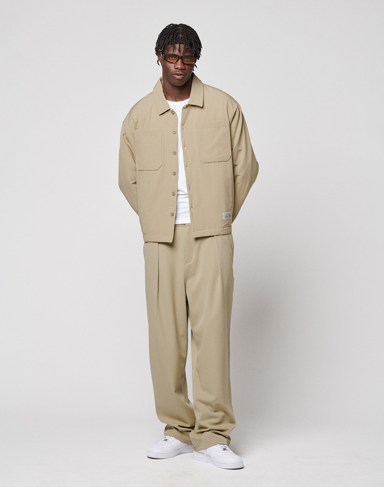 A man with short dreadlocks and glasses wears LIVE FAST DIE YOUNG Wide Casual Pants, a beige button-up overshirt, white t-shirt, and white sneakers, standing against a plain white background.