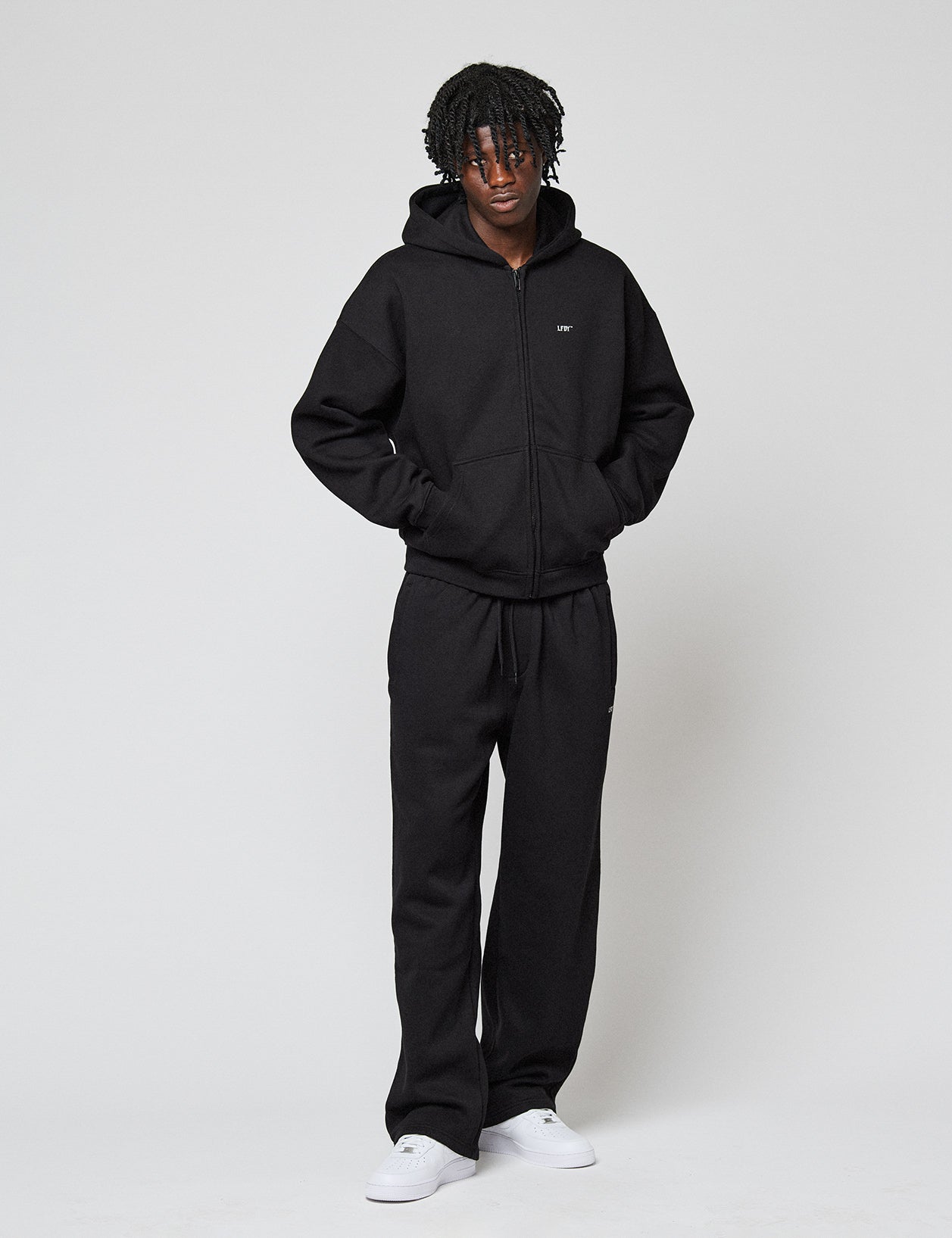 A person stands against a plain background wearing an Oversize Fit black zip-up hoodie, LIVE FAST DIE YOUNG Basic Wide Jogger black sweatpants with Elastikbund, and white sneakers, hands in pockets and a neutral expression.