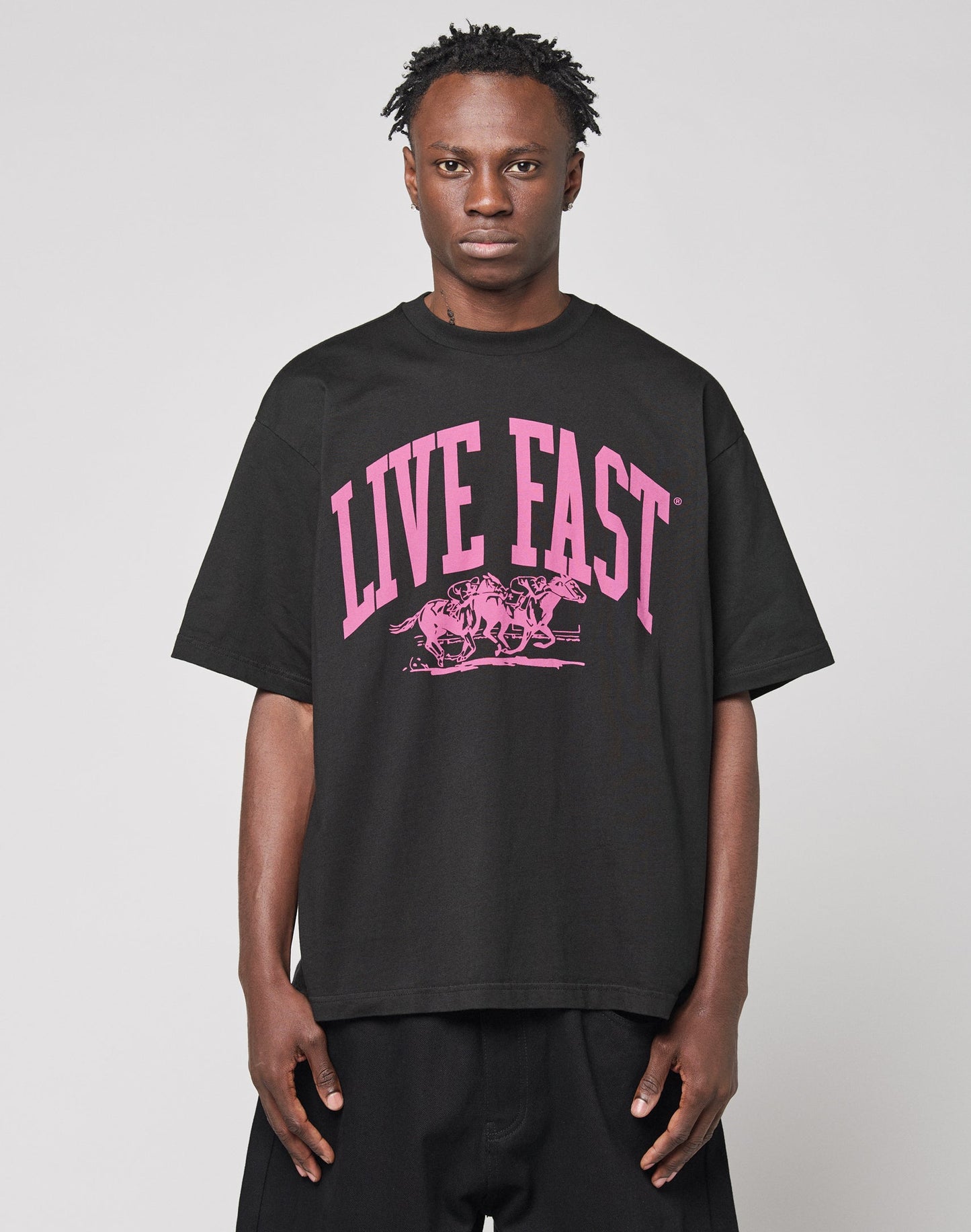 Wearing the Live Fast Stallion Tee by LIVE FAST DIE YOUNG—a black oversize 100% Baumwolle shirt with bold pink “LIVE FAST” frontprint above two horses—the person stands against a plain light gray background, facing forward neutrally.