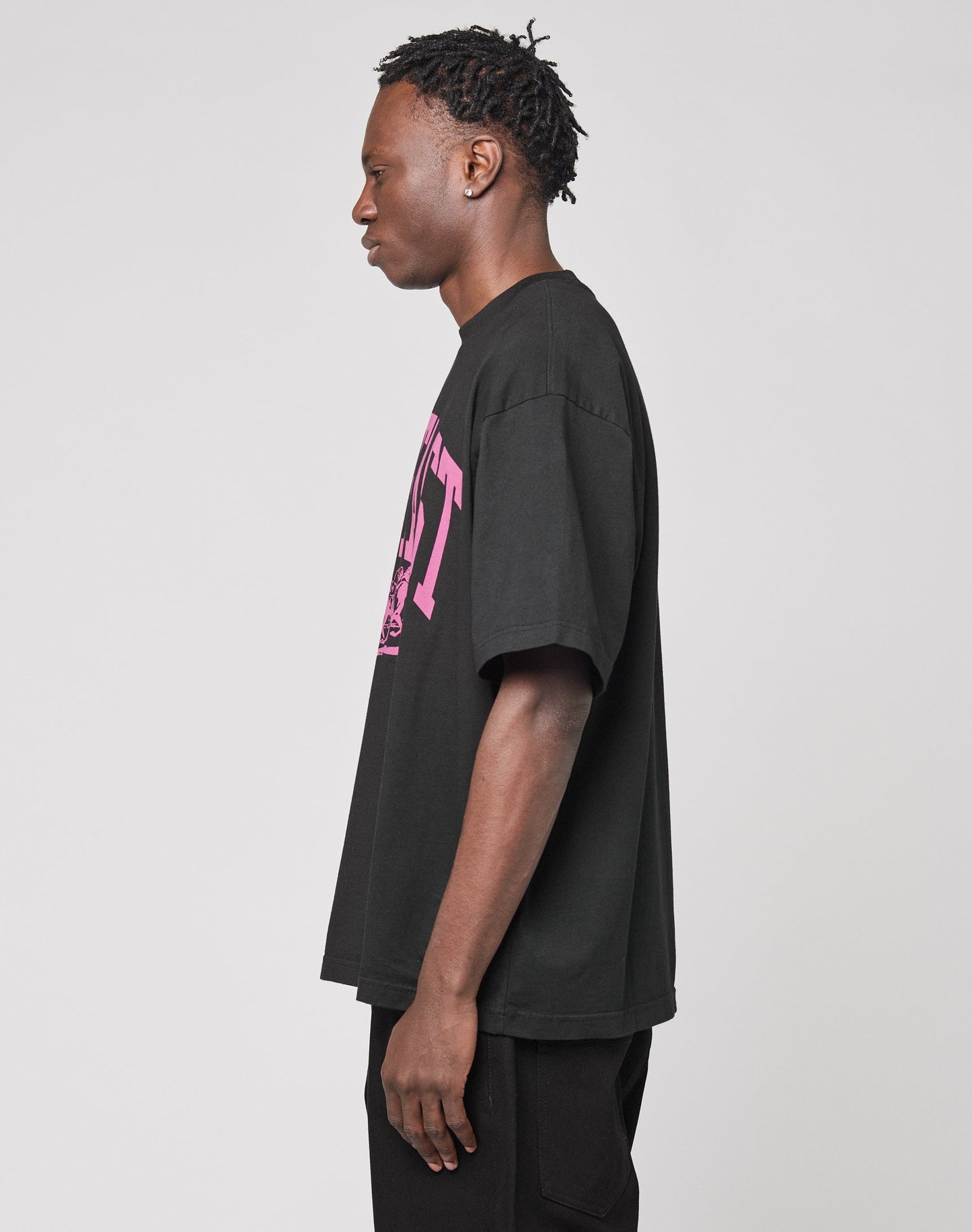 A young man with short twists stands in profile, wearing the LIVE FAST DIE YOUNG Live Fast Stallion Tee—an oversize black 100% Baumwolle T-shirt with a pink front print—paired with black pants, against a plain light gray background.