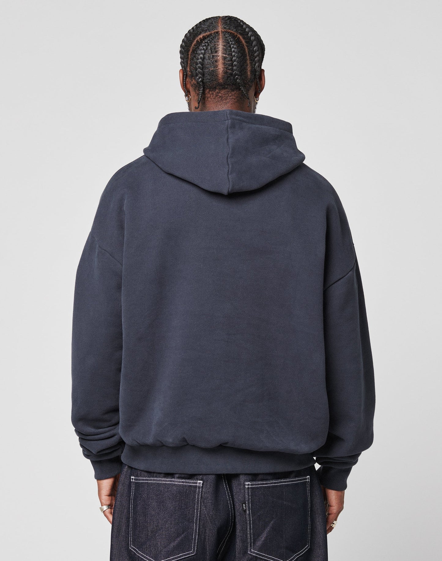 A person with braided hair is seen from behind, wearing a dark navy LFDY Script Hooded by LIVE FAST DIE YOUNG and dark denim jeans against a plain light gray background.