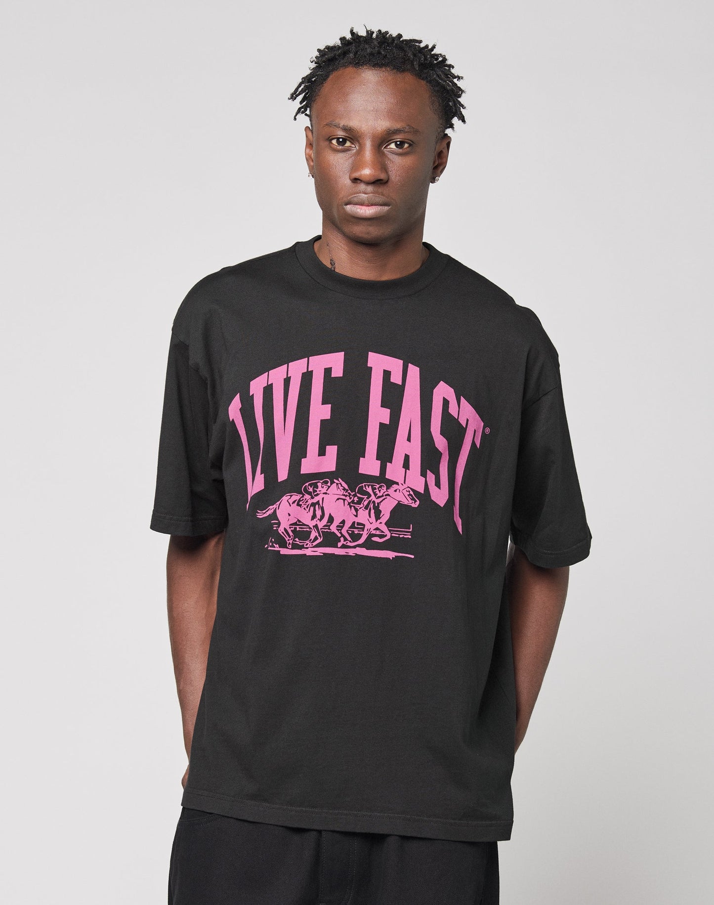 Someone wearing the Live Fast Stallion Tee by LIVE FAST DIE YOUNG, an oversize-fit black T-shirt featuring bold pink LIVE FAST text and a front print of three running animals, stands against a plain light background.