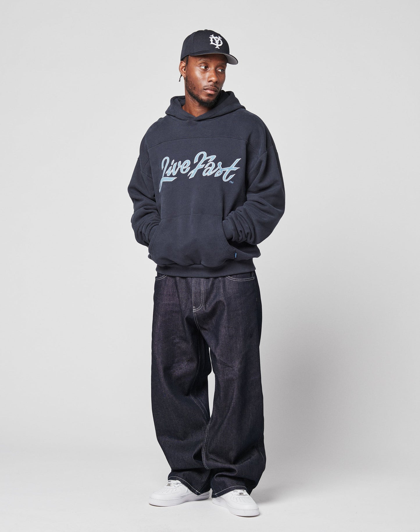 A man in a black cap, LIVE FAST DIE YOUNG LFDY Script Hooded oversize dark hoodie, baggy dark jeans, and white sneakers stands against a plain light gray background with his hands in his hoodie pockets.