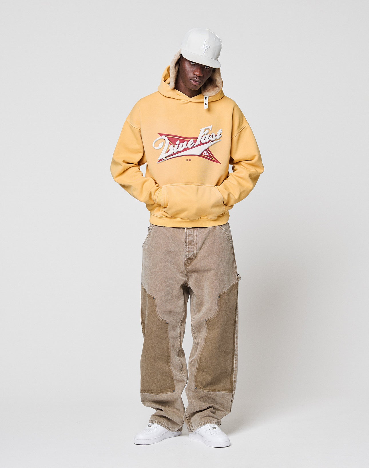A person in the yellow LFDY Corp. Hooded by LIVE FAST DIE YOUNG with a front print and spray effect, loose tan patched pants, white sneakers, and a white cap stands against a white background with hands in the hoodie pockets.