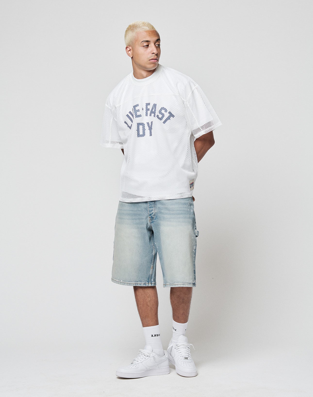 A man with short bleached blond hair wears the LFDY Boxy Jersey by LIVE FAST DIE YOUNG, featuring a bold frontprint, paired with light denim knee-length shorts, white socks, and white sneakers against a plain light background.
