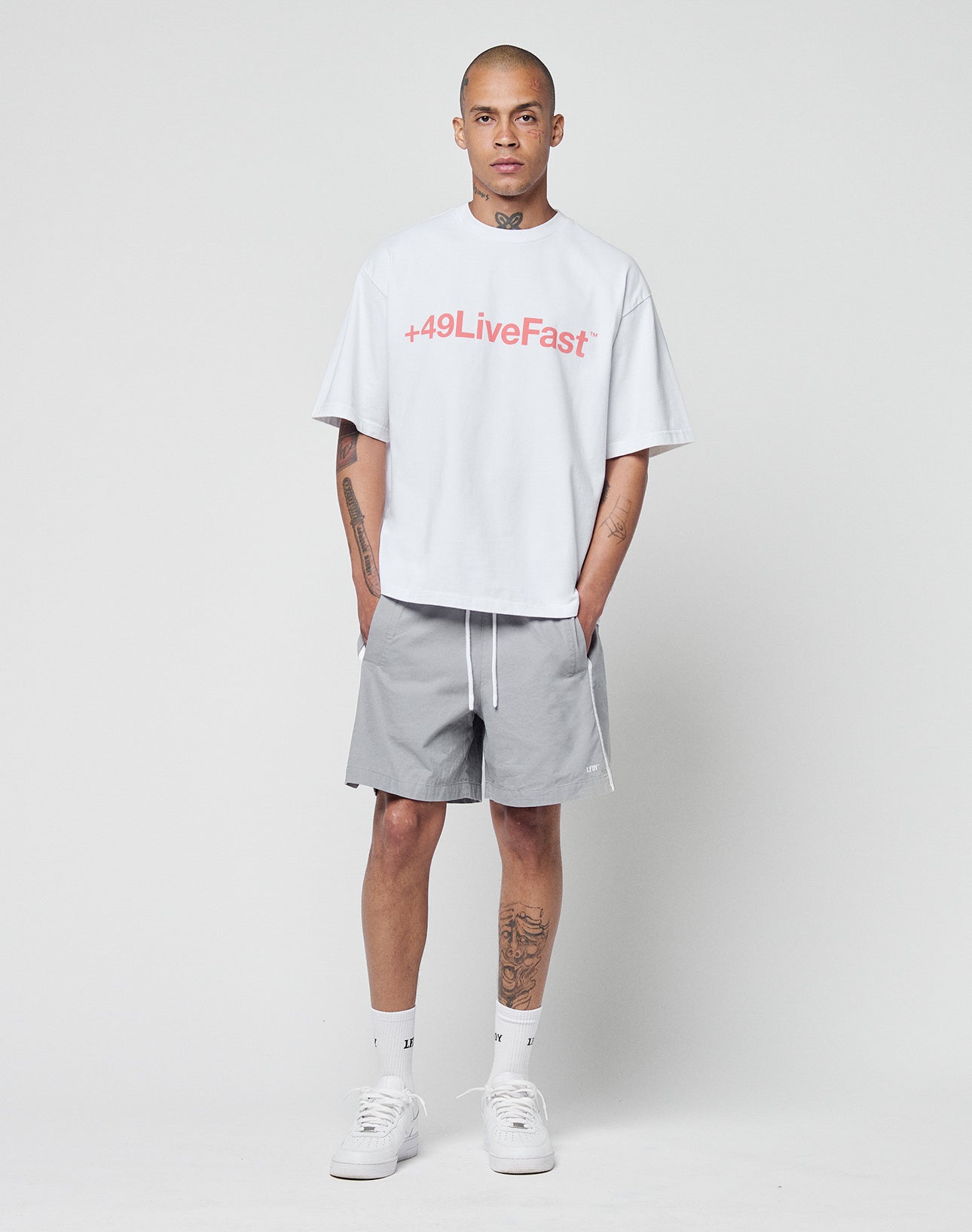 A person with tattoos and a shaved head stands against a plain background wearing a white oversized t-shirt with “+49LiveFast” logo print in pink, LIVE FAST DIE YOUNG Basic Cotton Shorts with elastischer Bund, white socks, and sneakers.