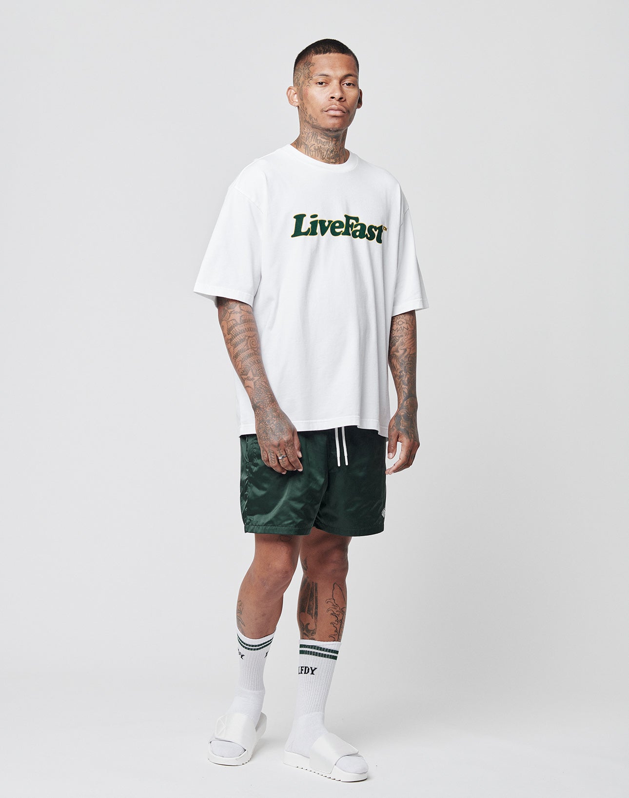 A man stands against a plain background wearing a white LIVE FAST DIE YOUNG Basic Tee in an oversize fit, dark green shorts, striped white socks, and white slide sandals. He has tattoos on his neck, arms, and legs.