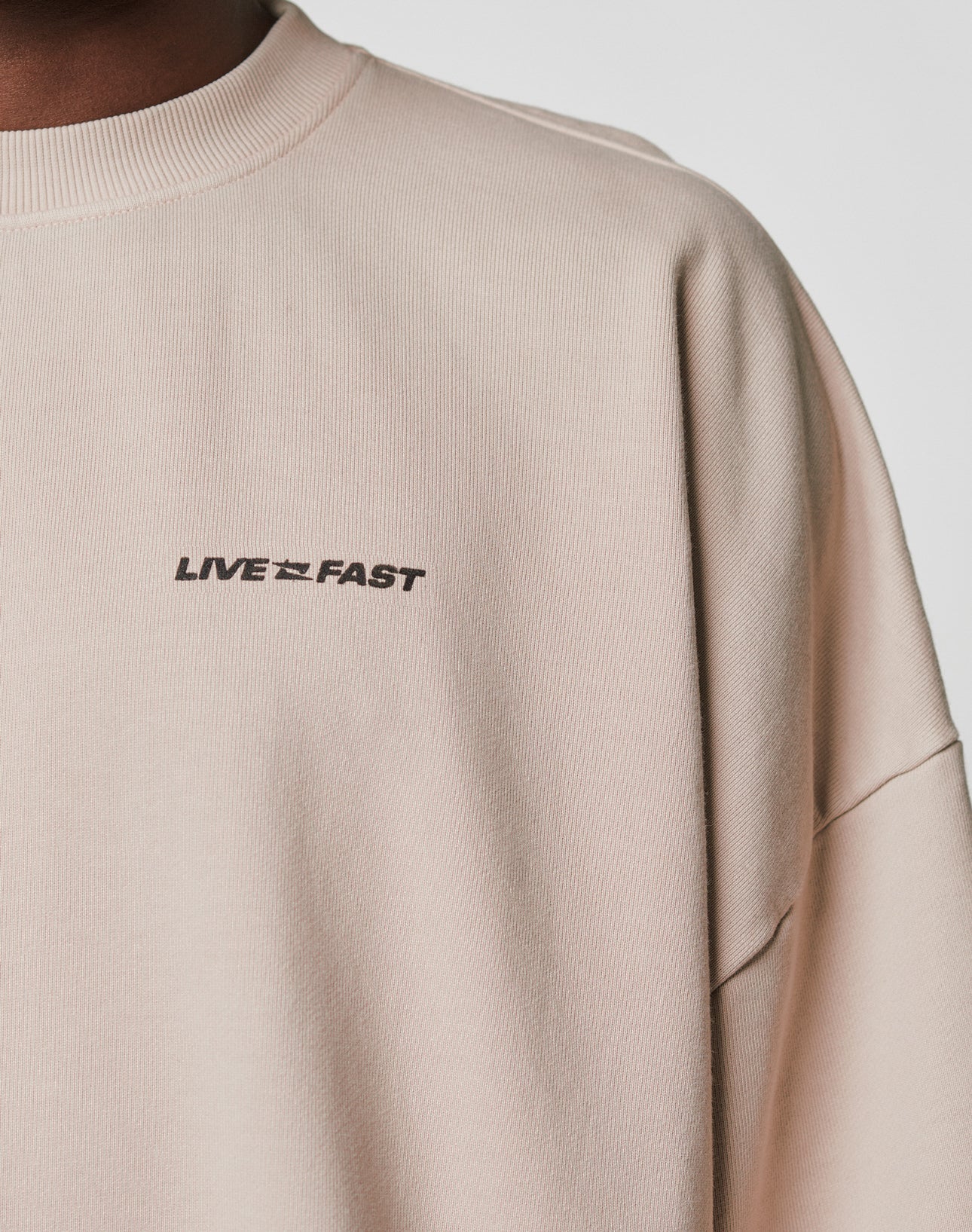 A close-up of the beige Live For It Sweater by LIVE FAST DIE YOUNG, featuring a distressed effect and black LIVE FAST print on the chest. Only part of the models neck and shoulder are shown.