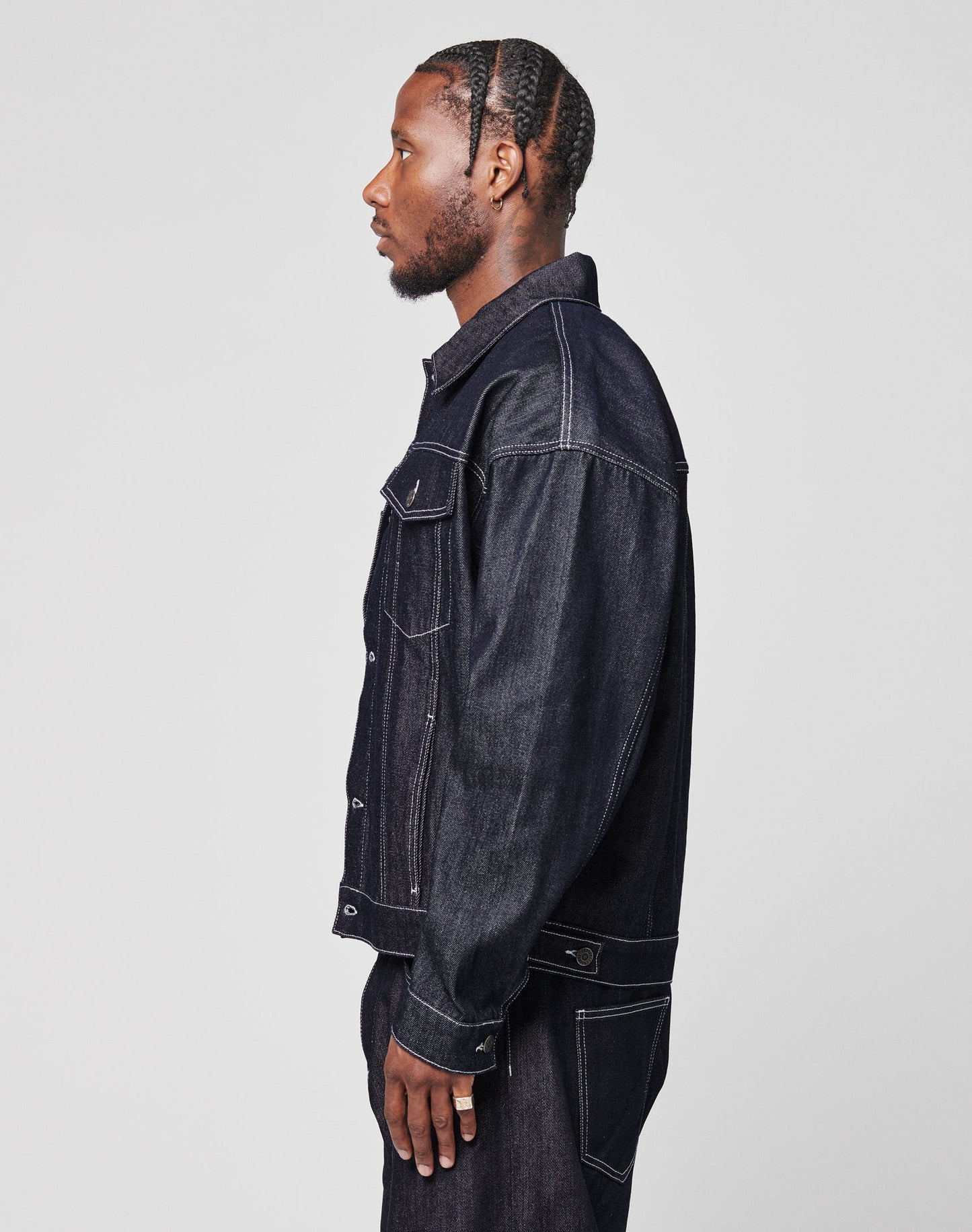 A man with braided hair stands in profile, wearing an oversize LFDY Denim Jacket and matching pants by LIVE FAST DIE YOUNG against a plain light gray background.