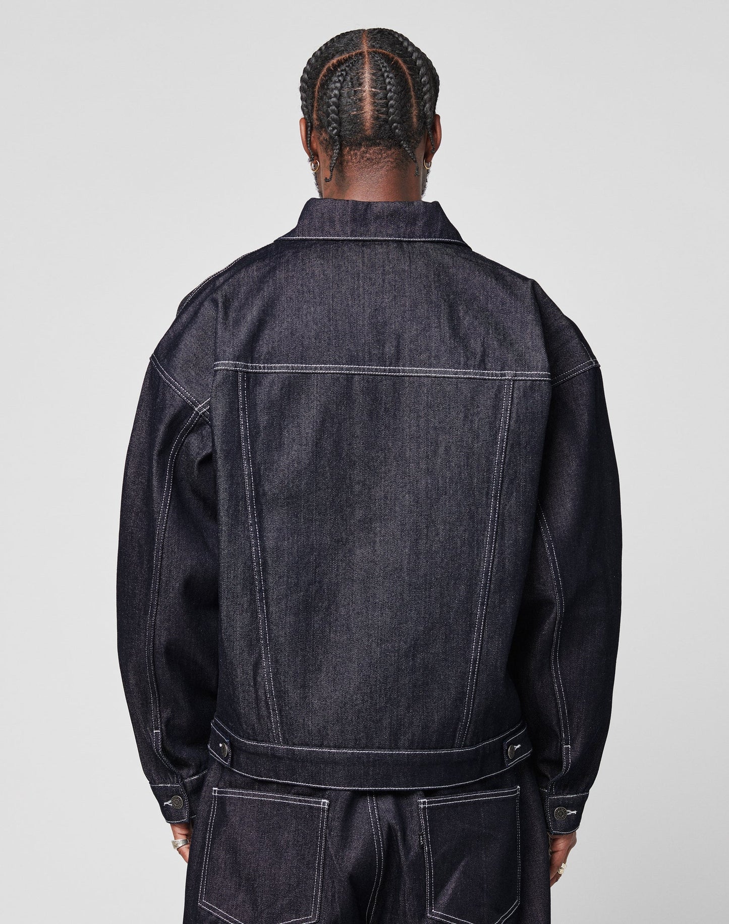 Person with braided hair facing away, wearing an oversize fit LFDY Denim Jacket by LIVE FAST DIE YOUNG and matching raw-denim jeans with visible white stitching, set against a plain light background.