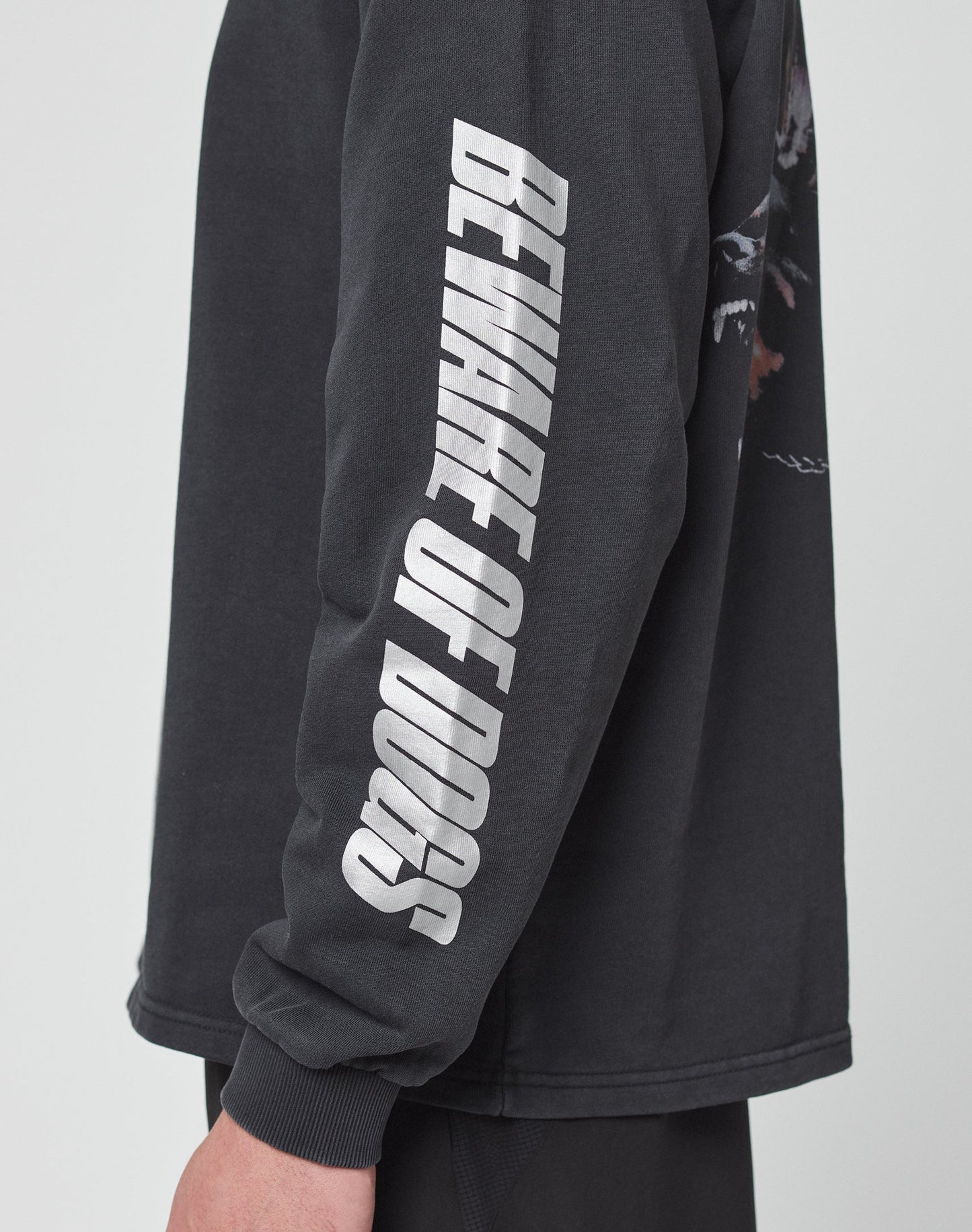 A person wears the LFDY Canine Longsleeve by LIVE FAST DIE YOUNG, an oversized black shirt with BEWARE OF DOGS in bold reflective print down the sleeve. Only their arm and part of their torso are visible.