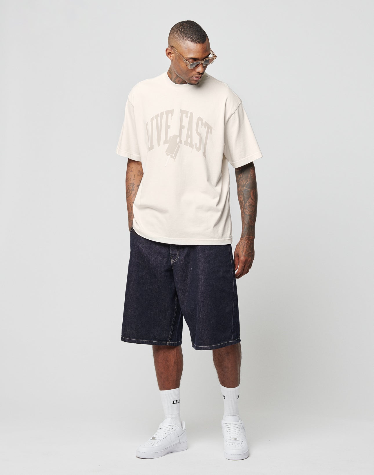 A man in glasses wears the LIVE FAST DIE YOUNG Live Fast Ice Tee (Oversize Fit), dark denim knee-length shorts, white socks, and white sneakers. He stands against a light gray background, looking down with his hands in his pockets.