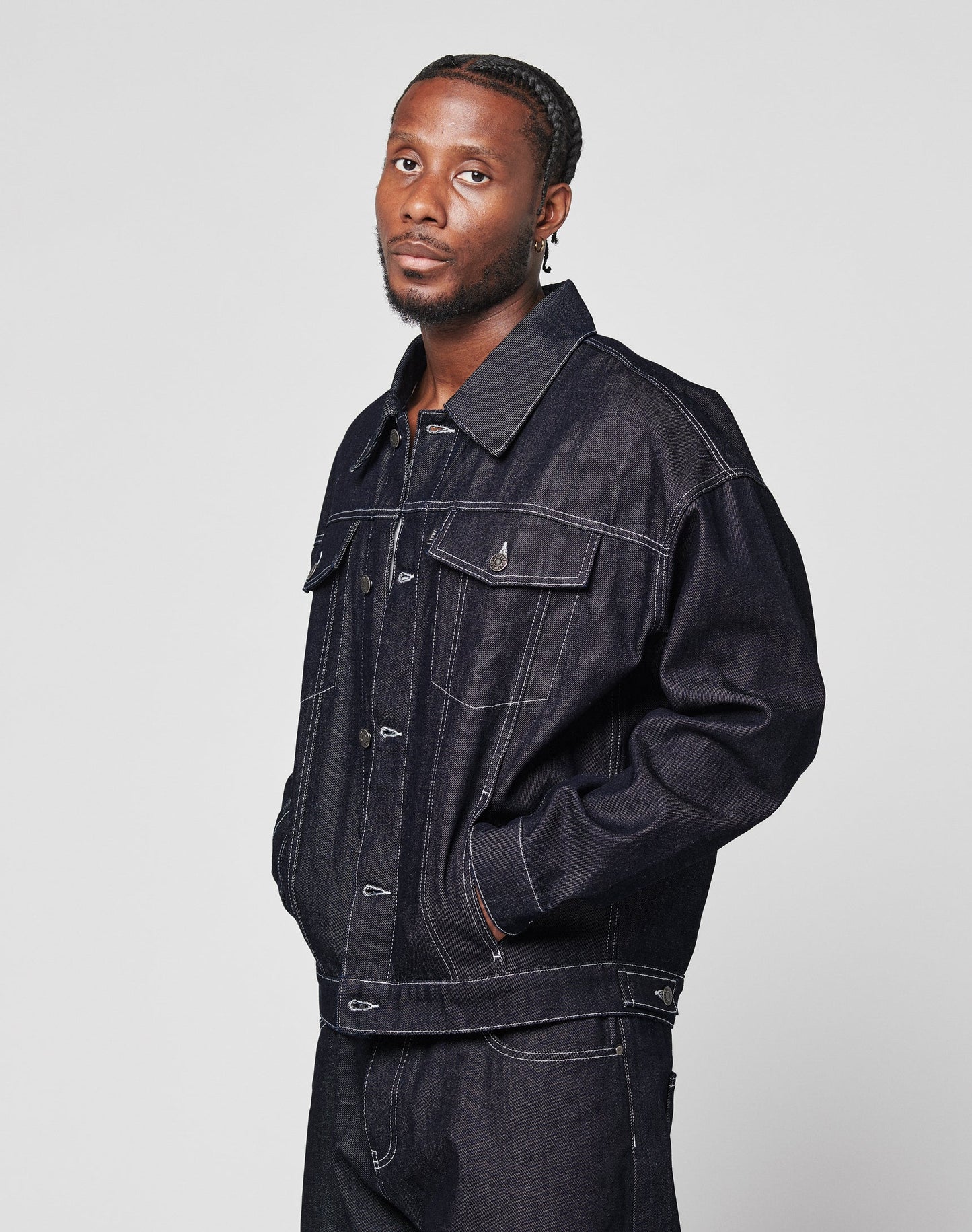 A man with braided hair and an earring, hands in pockets, wears the LIVE FAST DIE YOUNG LFDY Denim Jacket and matching 100% Baumwolle pants, calmly facing the camera against a plain light background.