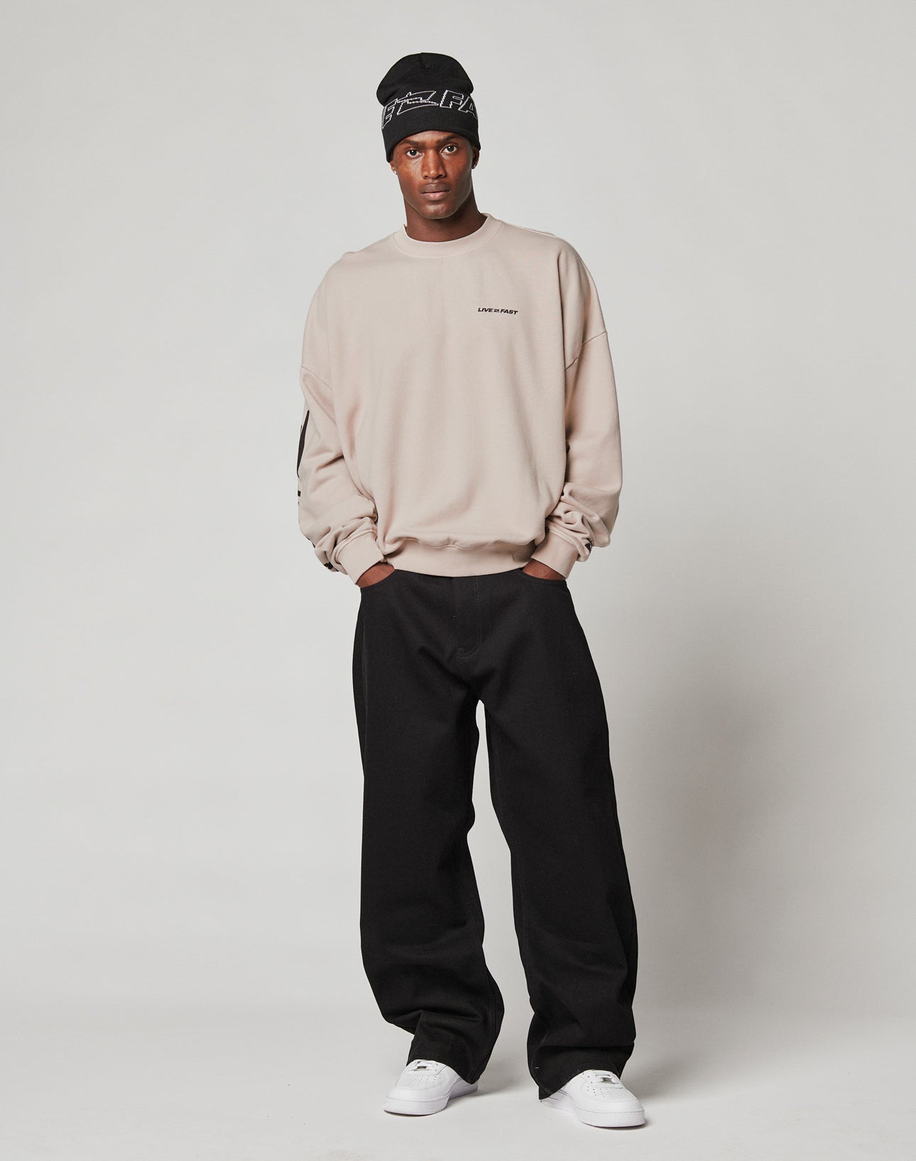 A man stands against a plain background wearing the beige Live For It Sweater by LIVE FAST DIE YOUNG, black baggy pants, white sneakers, and a black beanie. He has a neutral expression with his hands in his pockets.