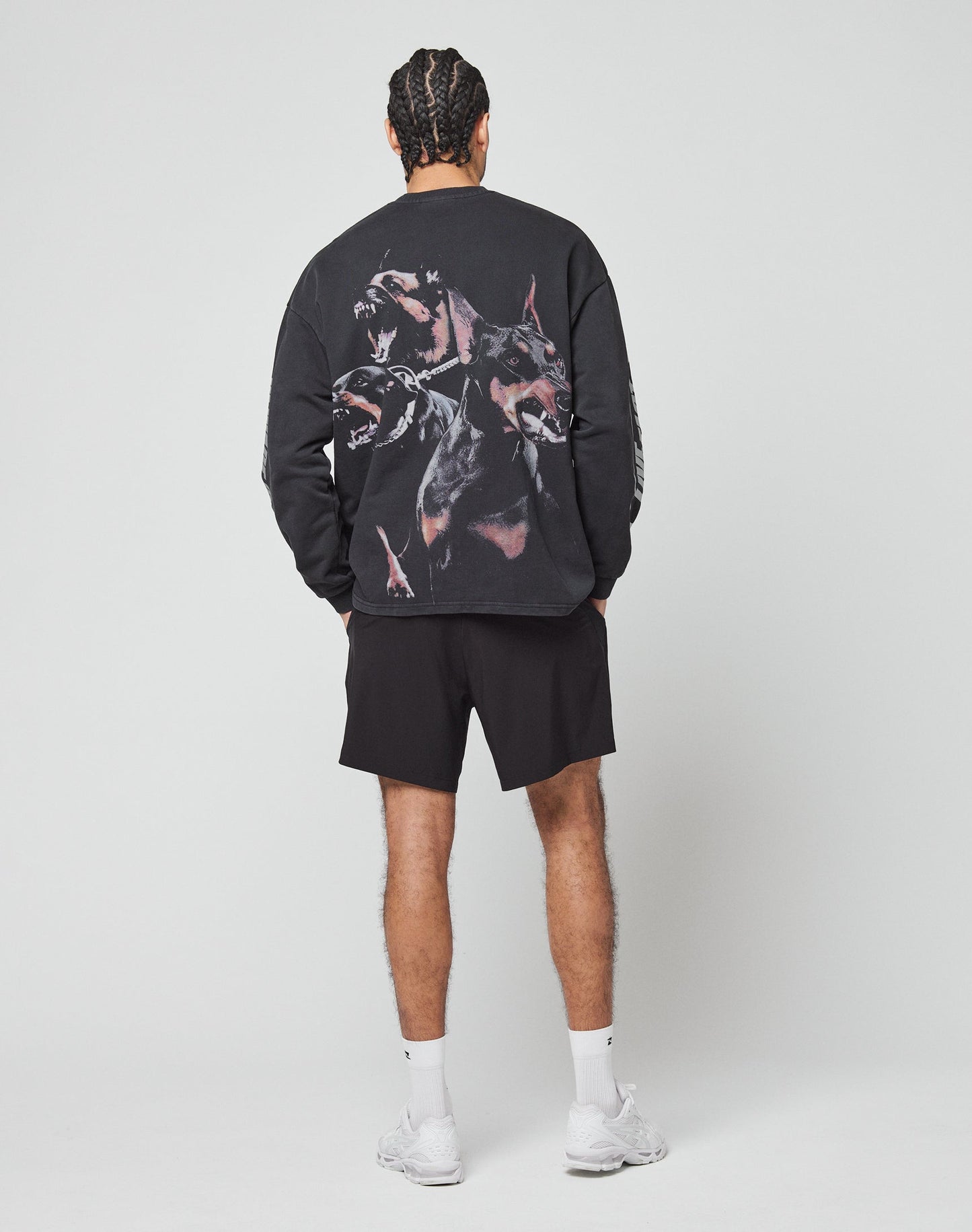 A person with braided hair faces away, wearing the LIVE FAST DIE YOUNG LFDY Canine Longsleeve—an oversized black sweatshirt featuring a large snarling dogs graphic on the back—styled with black shorts, white socks, and white sneakers against a plain light background.
