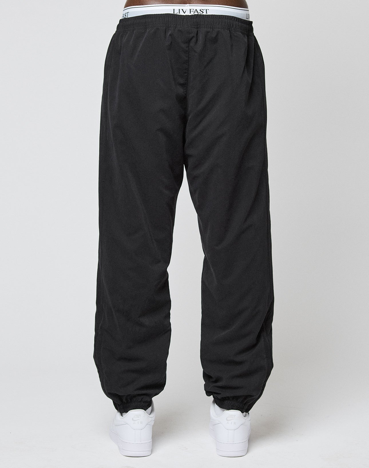 A person wearing loose-fitting, regular fit black LFDY Trackpants by LIVE FAST DIE YOUNG with elastic cuffs and waistband featuring LIV EAST stands against a plain light gray background, paired with white sneakers.