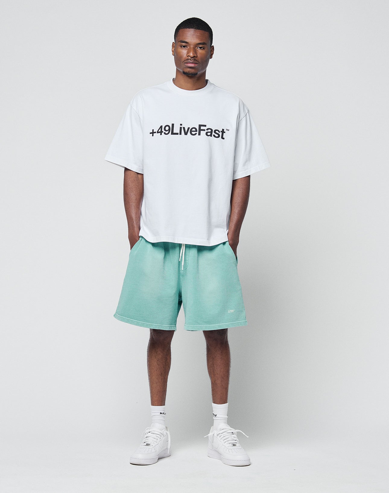 A man faces forward wearing a white, Regular Fit oversized T-shirt with +49LiveFast in black, LIVE FAST DIE YOUNG Basic Sweat Shorts in light green, white socks, and white sneakers against a plain light background.