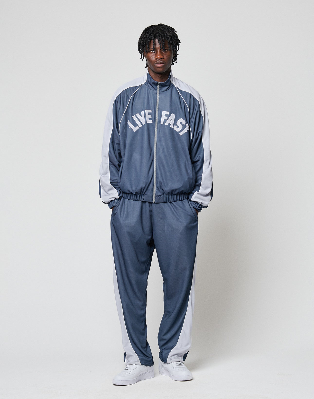 A person stands facing forward, wearing LIVE FAST DIE YOUNG LFDY DY Trackpants in blue and white polyester with an elastic waistband and LIVE FAST on the front, plus white sneakers, against a plain white background.