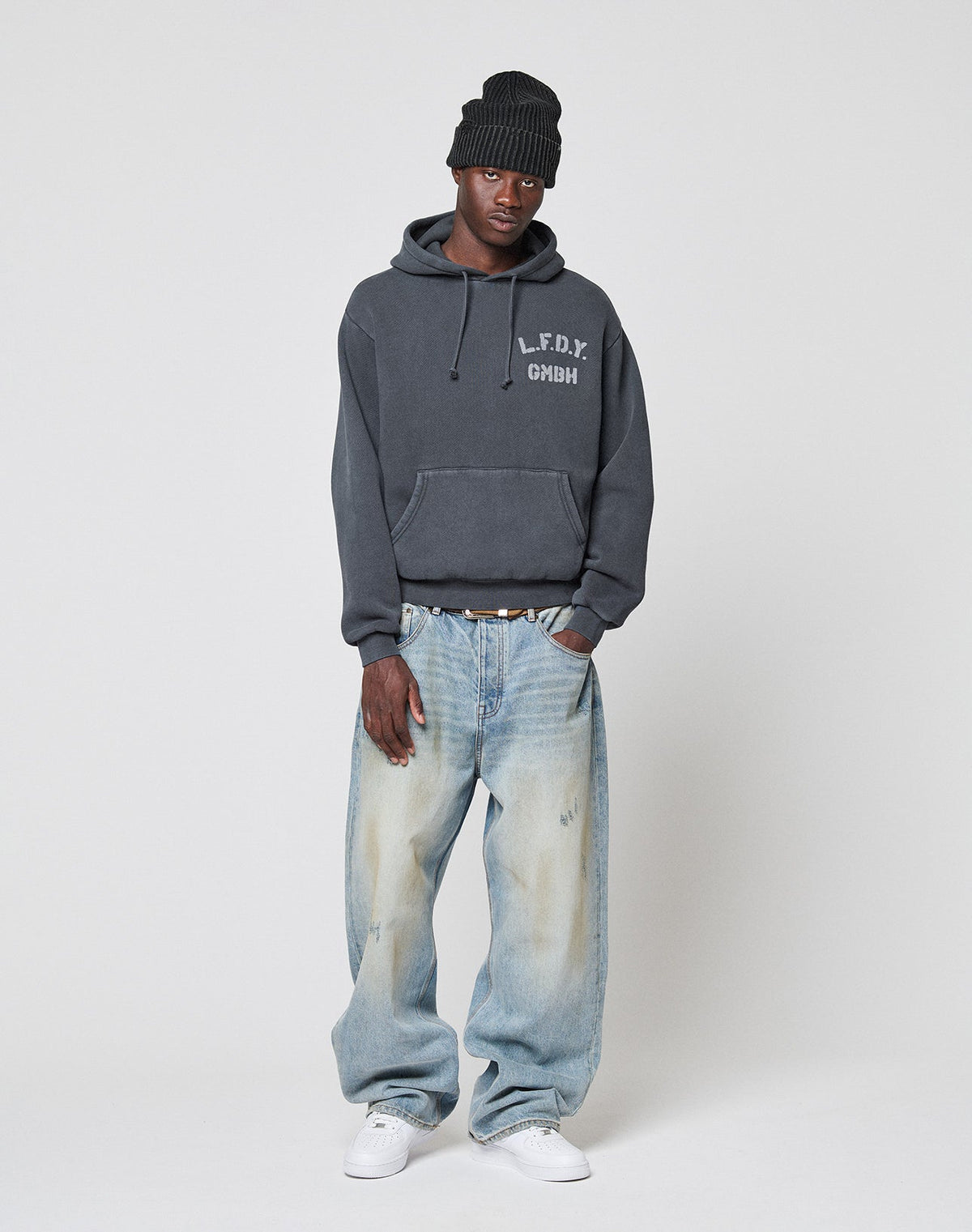 A person in a black beanie, LIVE FAST DIE YOUNG LFDY GmbH Hooded dark gray hoodie with Kapuze and text, baggy light blue oversize jeans, and white sneakers stands against a plain light background.
