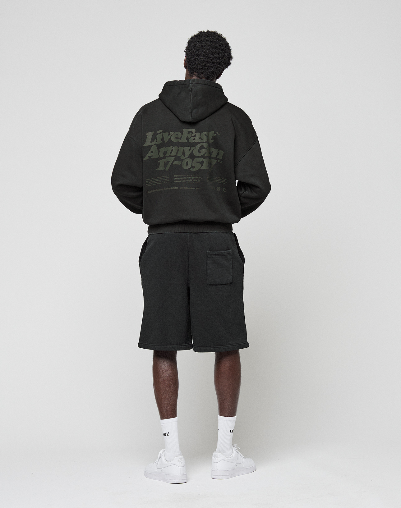 A person faces away, wearing the LIVE FAST DIE YOUNG Basic 360 Hooded black hoodie with "Live Fast Army Gn 17-051" back print, paired with black shorts, white socks, and white sneakers against a plain light background.