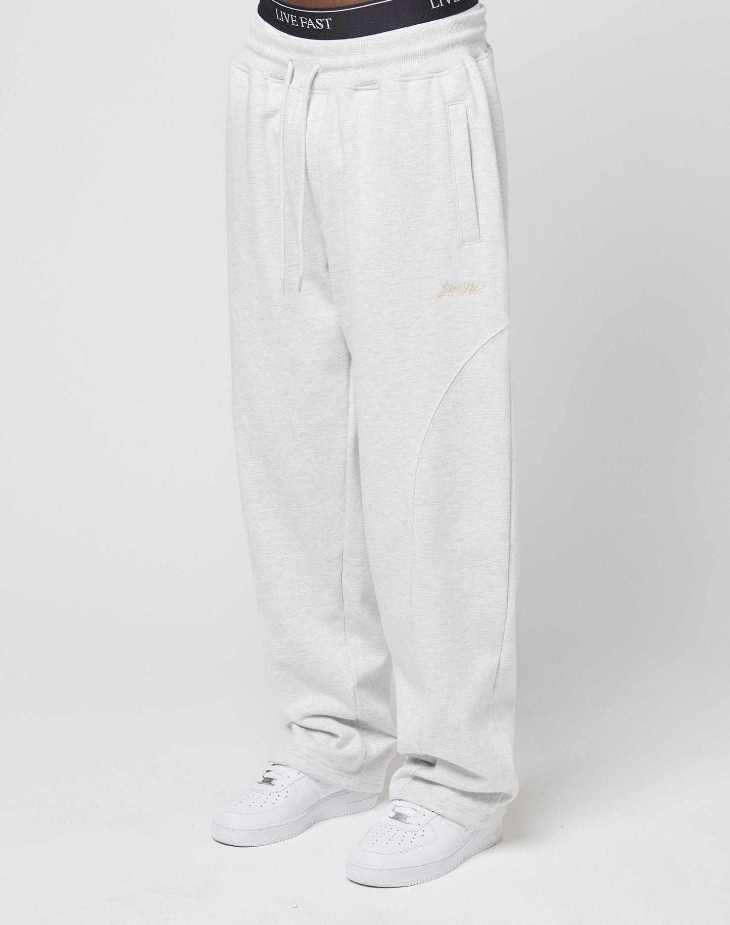 A person wears the LIVE FAST DIE YOUNG Basic Wide Jogger in light gray with an elastic drawstring waist and white sneakers. The black waistband above the pants reads LIVE FAST. Background is plain white.