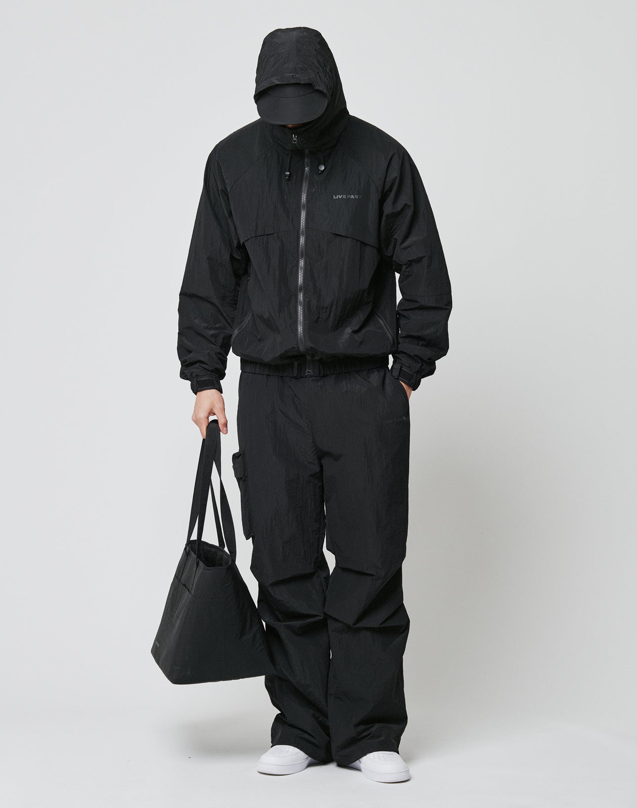 A person in an oversized Live Fast Nylon Jacket by LIVE FAST DIE YOUNG, black pants, black cap, and white shoes stands against a plain background, holding a black tote bag with silikon-print details. Their face is mostly hidden.